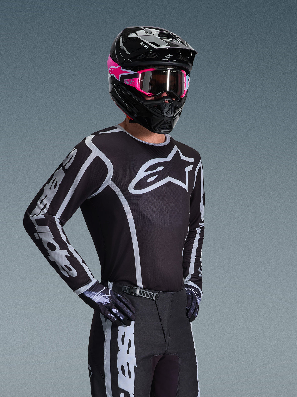 Motocross-kører iført Alpinestars 2026 Fluid Apex trøje, sort og grå med stort hvidt logo på brystet og geometrisk grafik på ærmerne, kombineret med sorte bukser, sorte handsker og en sort hjelm med lyserøde detaljer og lyserøde goggles