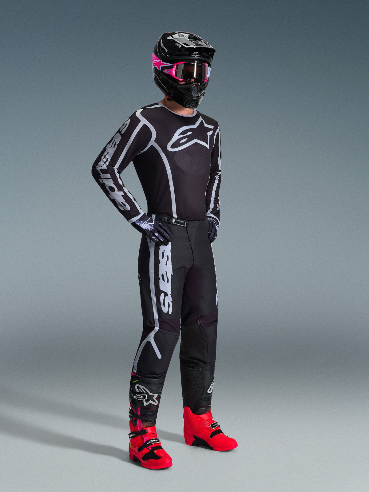 Motocross-kører iført 2026 Fluid Apex trøje, sort og grå, matchende bukser, sort hjelm med lyserøde goggles og knaldrøde støvler, markant hvidt Alpinestars-branding på bryst og ærmer, koordineret professionelt off-road raceroutfit.