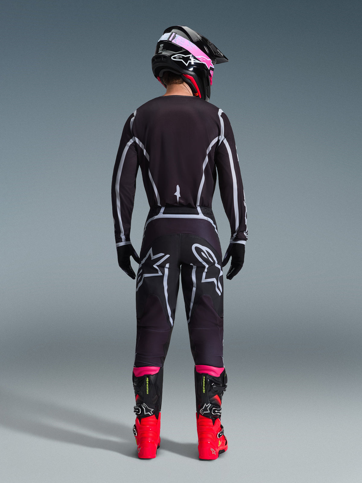 Motocross-kører set bagfra iført Alpinestars 2026 Fluid Apex trøje og matchende bukser i sort og grå, med hvide geometriske designlinjer og stjernelogoer på bagdelen, suppleret med en sort og pink hjelm og røde og sorte racingstøvler.