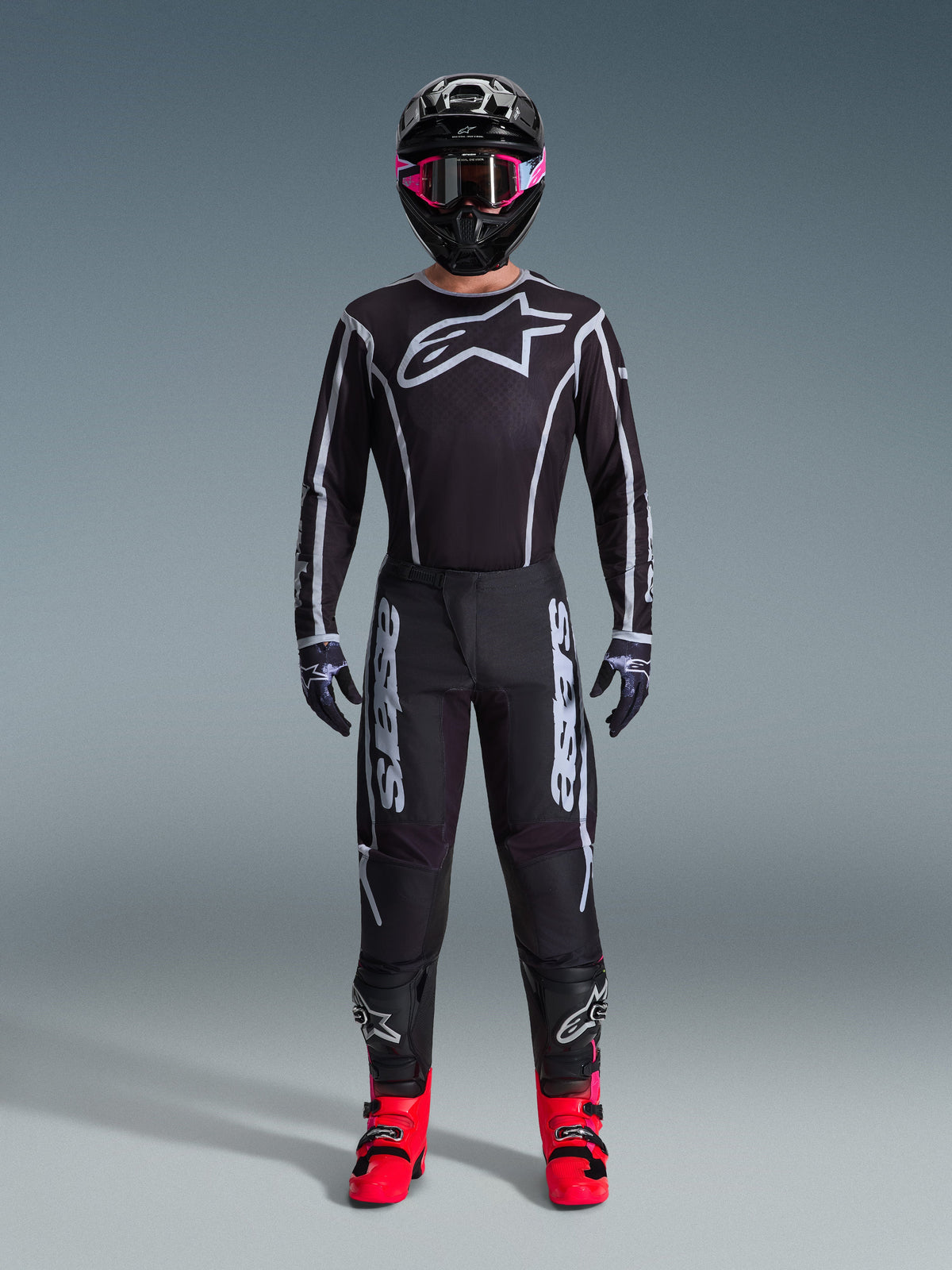 Motocrosskører iført Alpinestars 2026 Fluid Apex Jersey, sort og grå, stort hvidt Astars-logo på brystet, hvide geometriske linjer, matchende sorte bukser, sort hjelm med lyserøde goggles og panglyserøde støvler, komplet motocross-raceroutfit