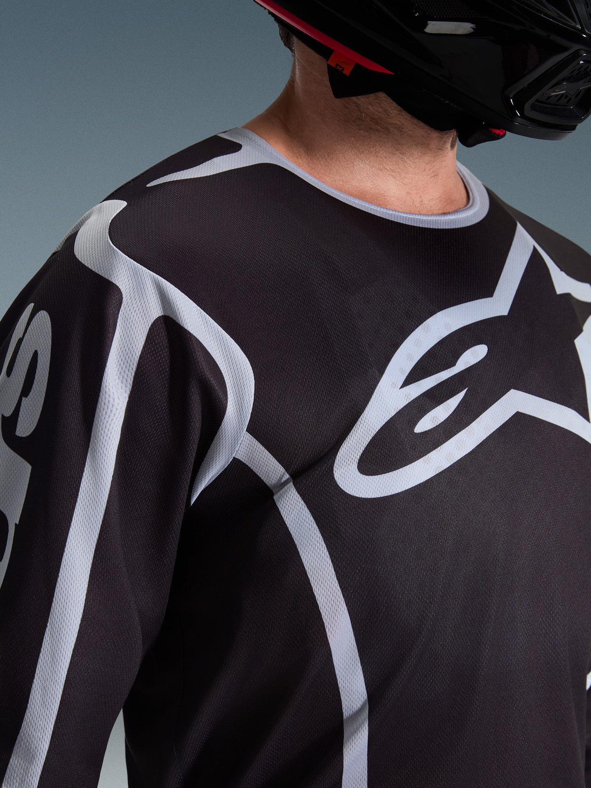Nærbillede af en kører iført Alpinestars 2026 Fluid Apex Jersey i sort og grå, der fremviser det store hvide Astars-logo på brystet og den tekniske mesh-konstruktion, kombineret med en sort motocrosshjelm.