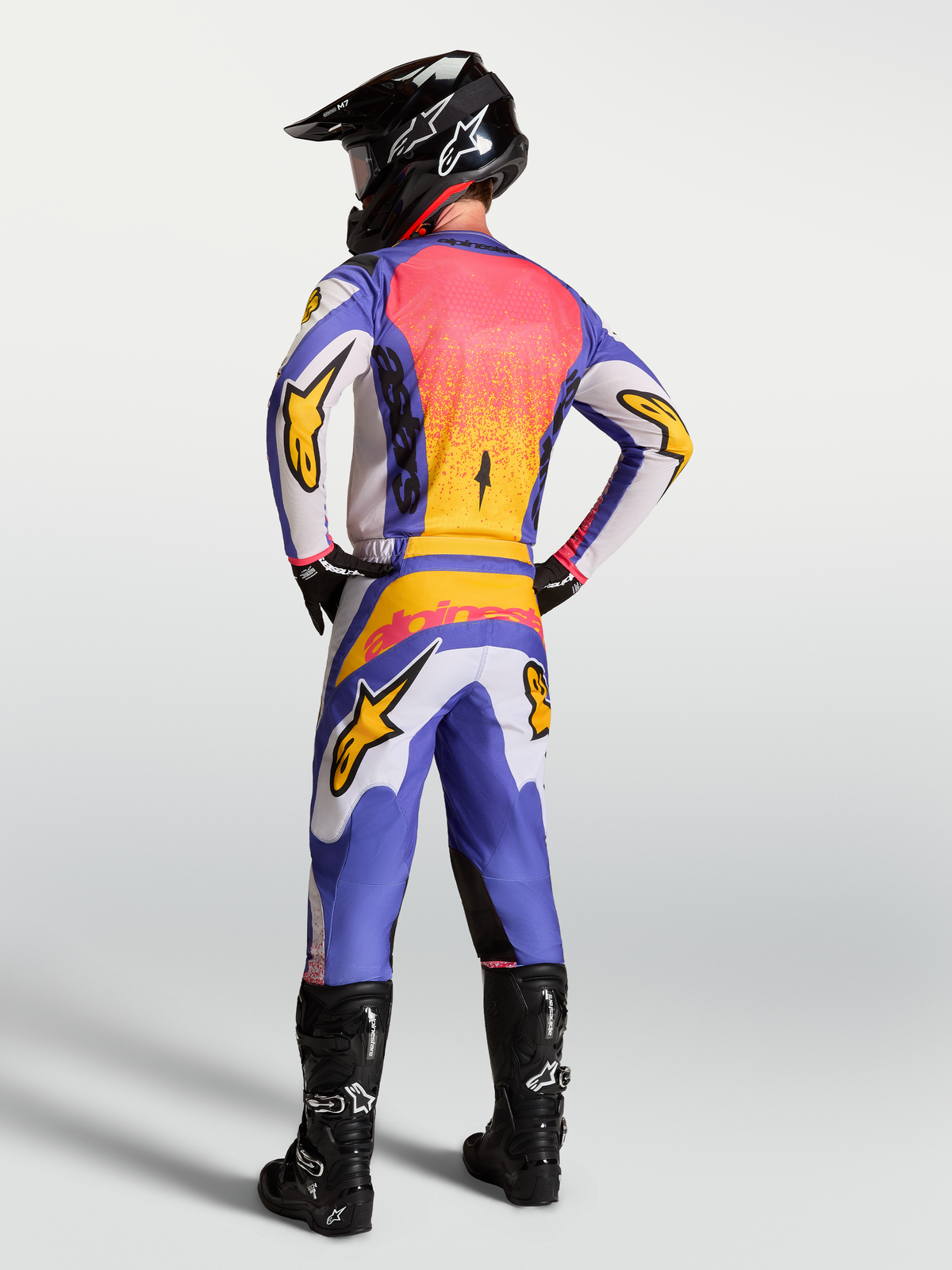 Alpinestars Fluid Drag Jersey in lilac and blue with gold accents, shown on a motocross rider from behind in full gear.