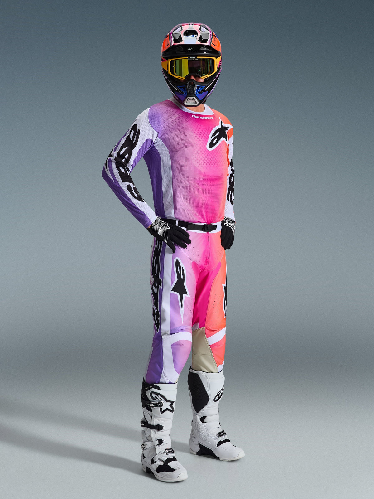 Alpinestars 2026 Racer Air Portl bukser, motocross-kører iført komplet racerudstyr, lyserødt, lilla og orange flerfarvet design med hvide detaljer, matchende langærmet trøje, sort hjelm med gule goggles og hvide støvler, professionelt udstyr til terrænkørsel