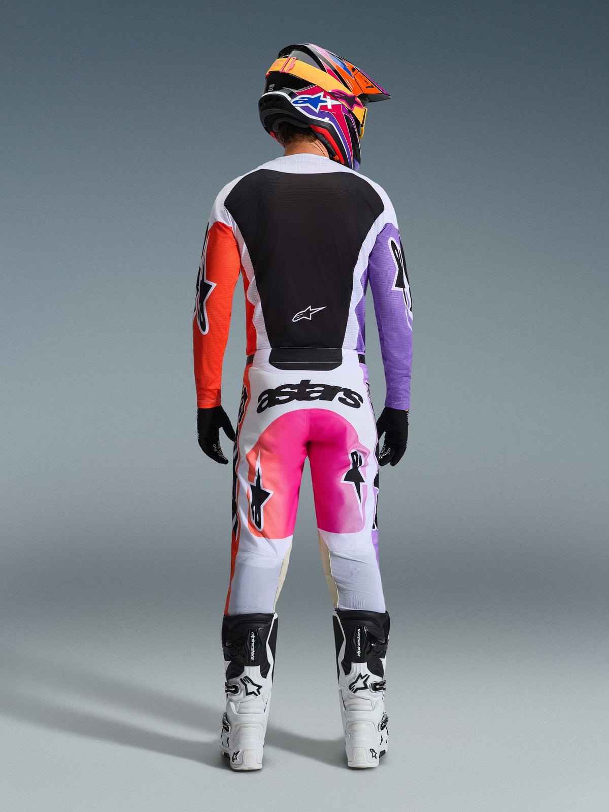 Set bagfra ses en motocrosskører iført Alpinestars 2026 Racer Air Portl-bukser i Multicolor, med hvidt stof, pink og orange gradientpaneler og sort branding, stylet med en matchende sort jersey med lilla og orange ærmer, hvide racestøvler og en livlig, flerfarvet hjelm.