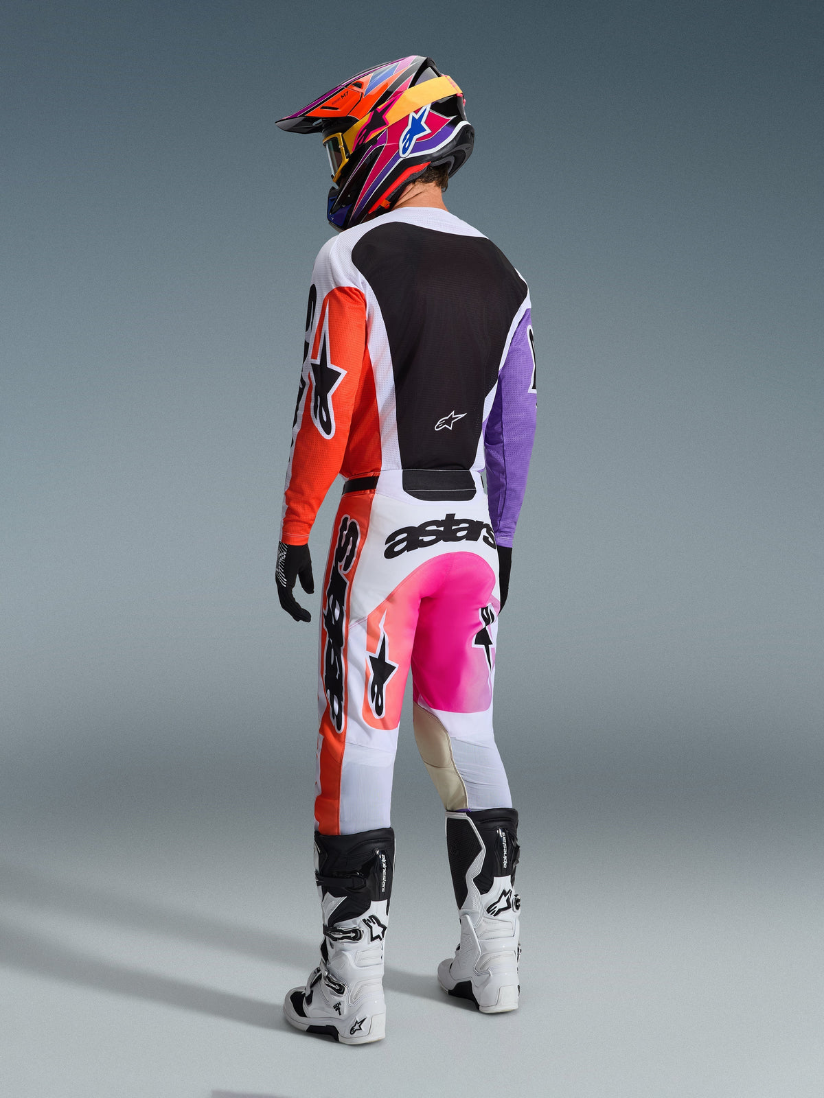 Motocrosskører set bagfra iført Alpinestars 2026 Racer Air Portl-bukser i Multicolor, med hvide, orange og lilla paneler, neon-lyserøde detaljer og markant sort branding, kombineret med en matchende trøje, farverig mønstret hjelm og hvide og sorte støvler.