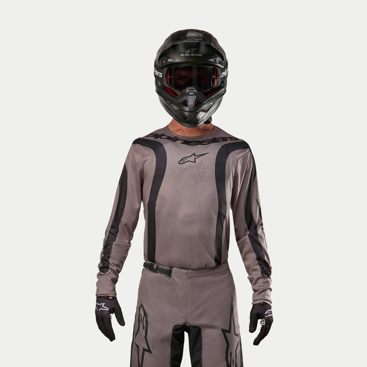 Motocrossrytter iført Alpinestars 2024 Fluid Lurv Jersey i Mud Black, et jordnært gråbrunt stof med sorte grafiske striber og stjernelogo på brystet, kombineret med en sort hjelm, røde goggles og sorte handsker for et koordineret off-road racinglook