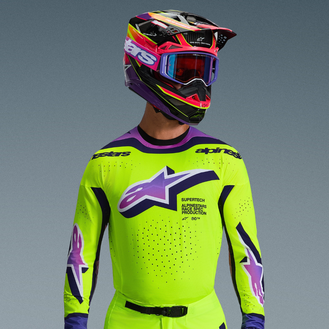 Alpinestars 2026 Supertech Vista-udstyr, motocrosskører i neongul trøje med lilla og sorte detaljer, stort lilla gradient-logo på brystet, matchende flerfarvet hjelm og lilla crossbriller, laserskåret ventilation, professionel atletisk pasform