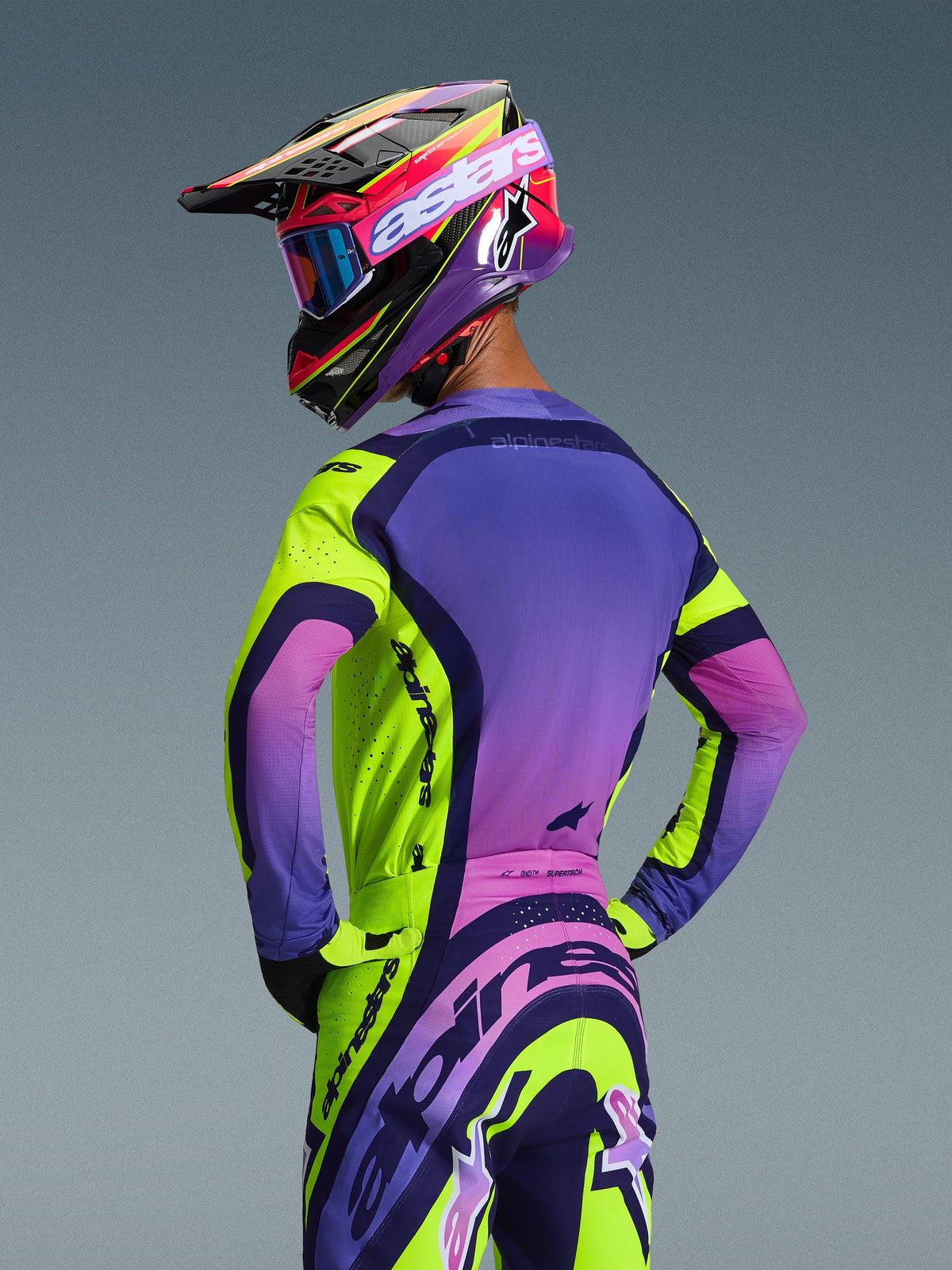 Motocrosskører set bagfra iført Alpinestars 2026 Supertech Vista-udstyrssæt, gul, lilla og sort trøje og bukser med et levende gradientdesign, mesh-ventilation og markant Alpinestars-branding, koordineret med en lilla hjelm og goggles.