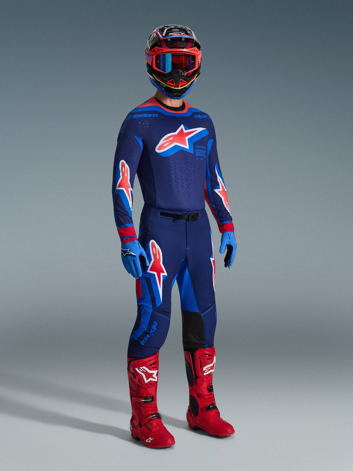 Motocross-kører iført komplet racerudstyr, 2026 Supertech Vista Jersey i mørkeblå med Hot Coral og koboltblå detaljer, matchende motocross-bukser, flerfarvet hjelm med røde goggles, klare røde støvler, blå handsker, koordineret outfit til terrænkørsel