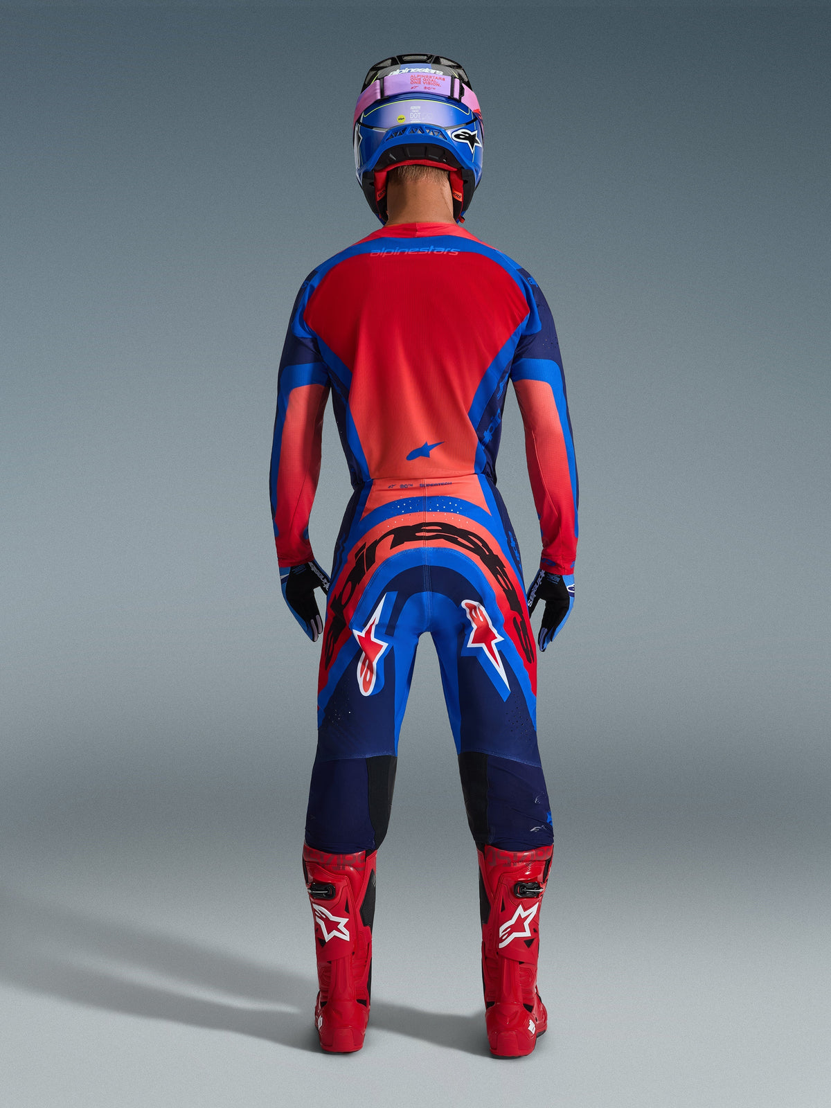 Alpinestars 2026 Supertech Vista Gear Set, motocrosskører set bagfra, Dark Navy, Hot Coral og Cobalt, klar rød ryg på trøjen, markant branding på bukserne, matchende røde støvler og blå og lyserød hjelm, professionelt motocross-udstyr