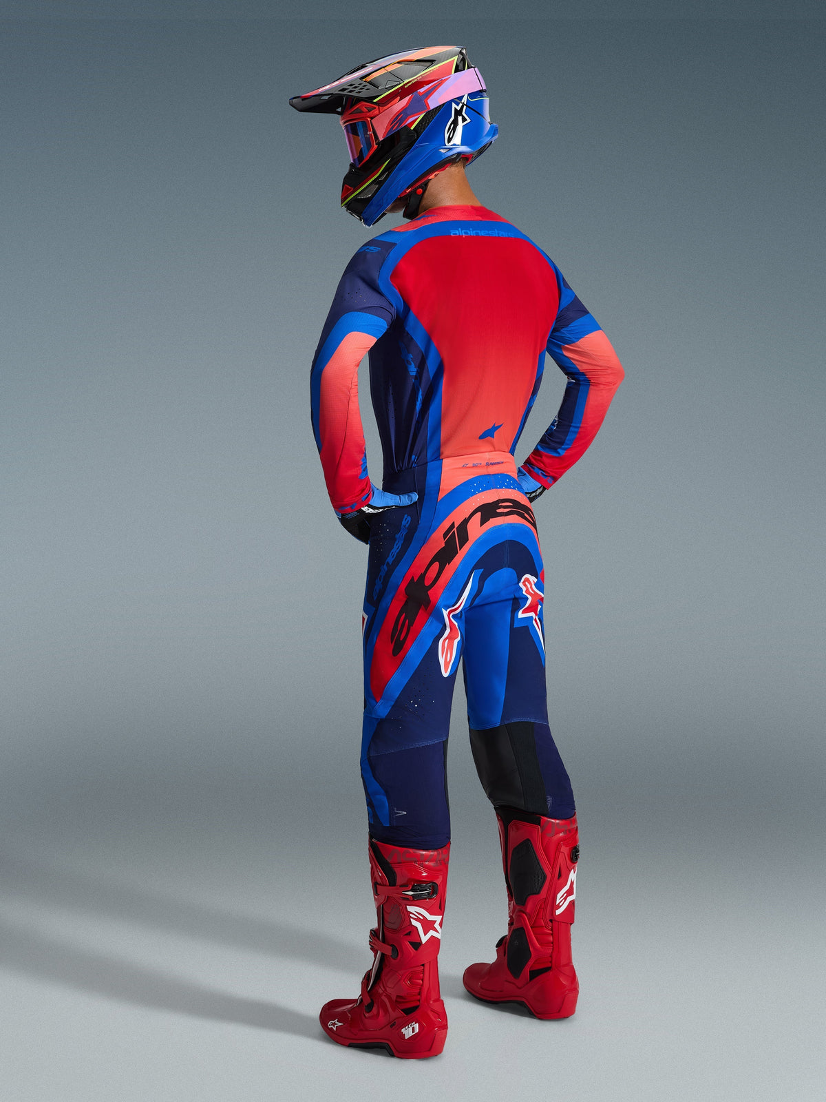 Alpinestars 2026 Supertech Vista udstyrssæt, set bagfra af en motocrosskører i Dark Navy, Hot Coral og Cobalt trøje og bukser, kombineret med en flerfarvet hjelm og knaldrøde motocrossstøvler, professionelt terrænkørselsudstyr.