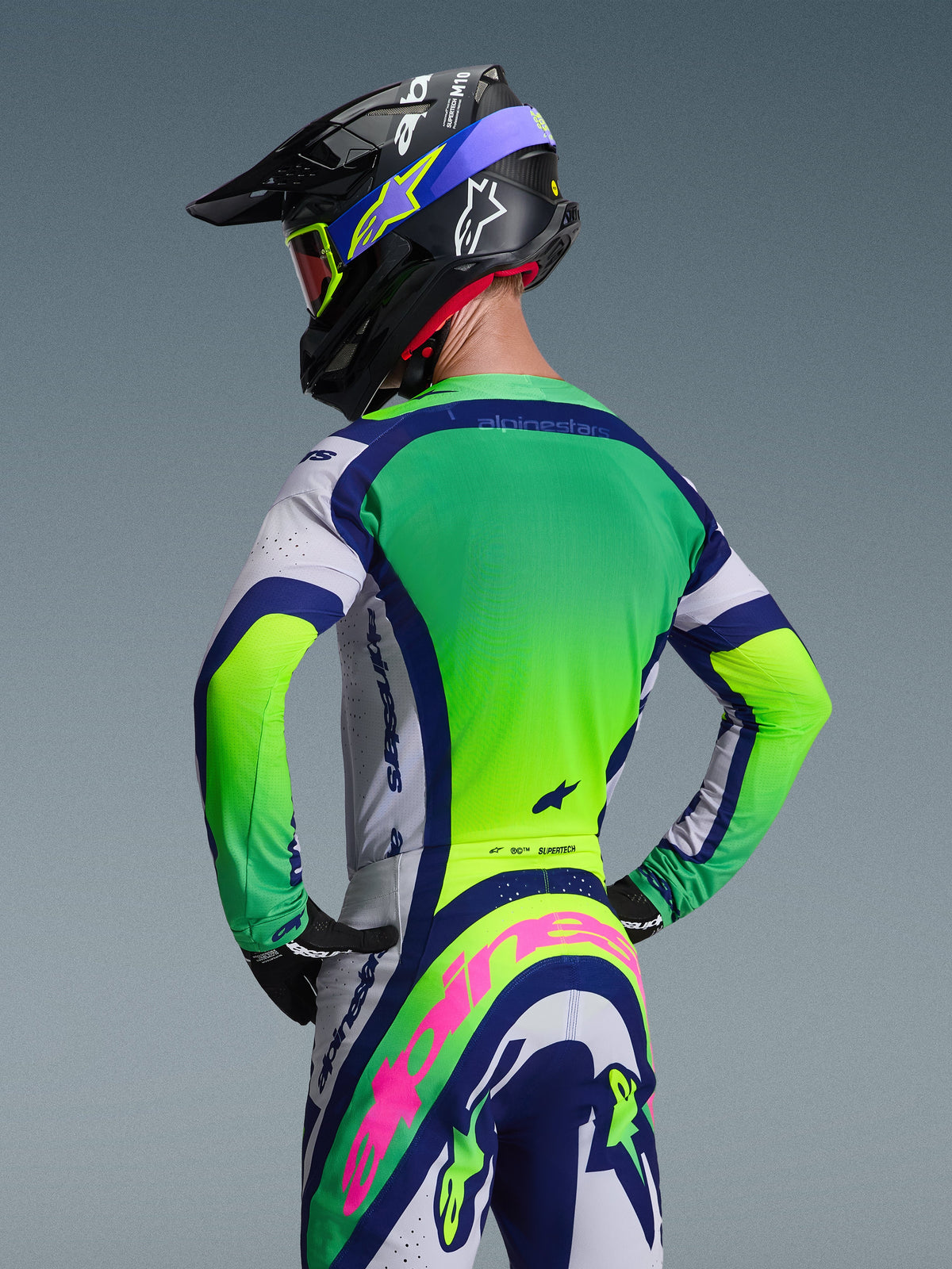 Alpinestars 2026 Supertech Vista Gear Set, motocross-udstyr set bagfra, grøn og hvid jersey med navyblå detaljer, matchende bukser med et markant lyserødt Alpinestars-logo, sort racerhjelm med blå grafik og røde goggles, neutral grå baggrund
