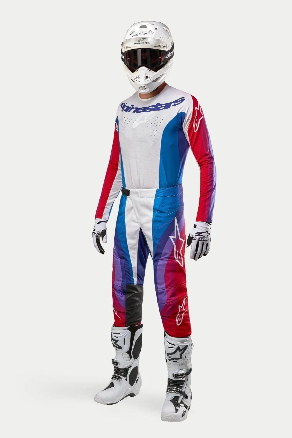 Motocross-kører i komplet Alpinestars-udstyr, 2024 Techstar Pneuma bukser i blå, rød og hvid, matchende langærmet jersey, hvid hjelm med mørke goggles og hvide og sorte støvler, koordineret outfit til terrænkørsel.