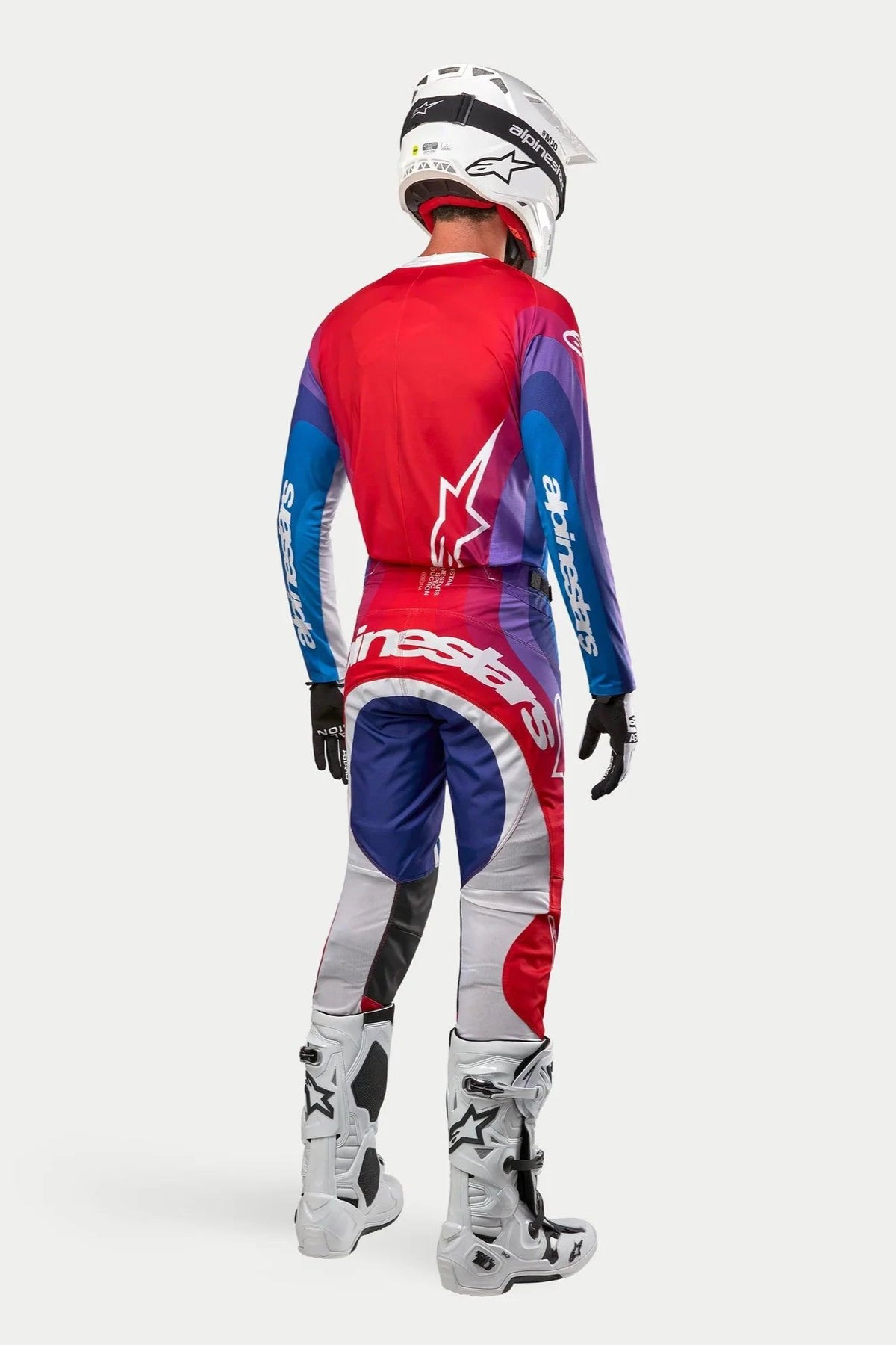 Motocrosskører set bagfra iført Alpinestars 2024 Techstar Pneuma trøje og bukser, Blu Mars Red White-farvekombination med røde, blå og lilla gradueringer og hvid branding, kombineret med hvide støvler og hvid hjelm, koordineret off-road racerudstyr.