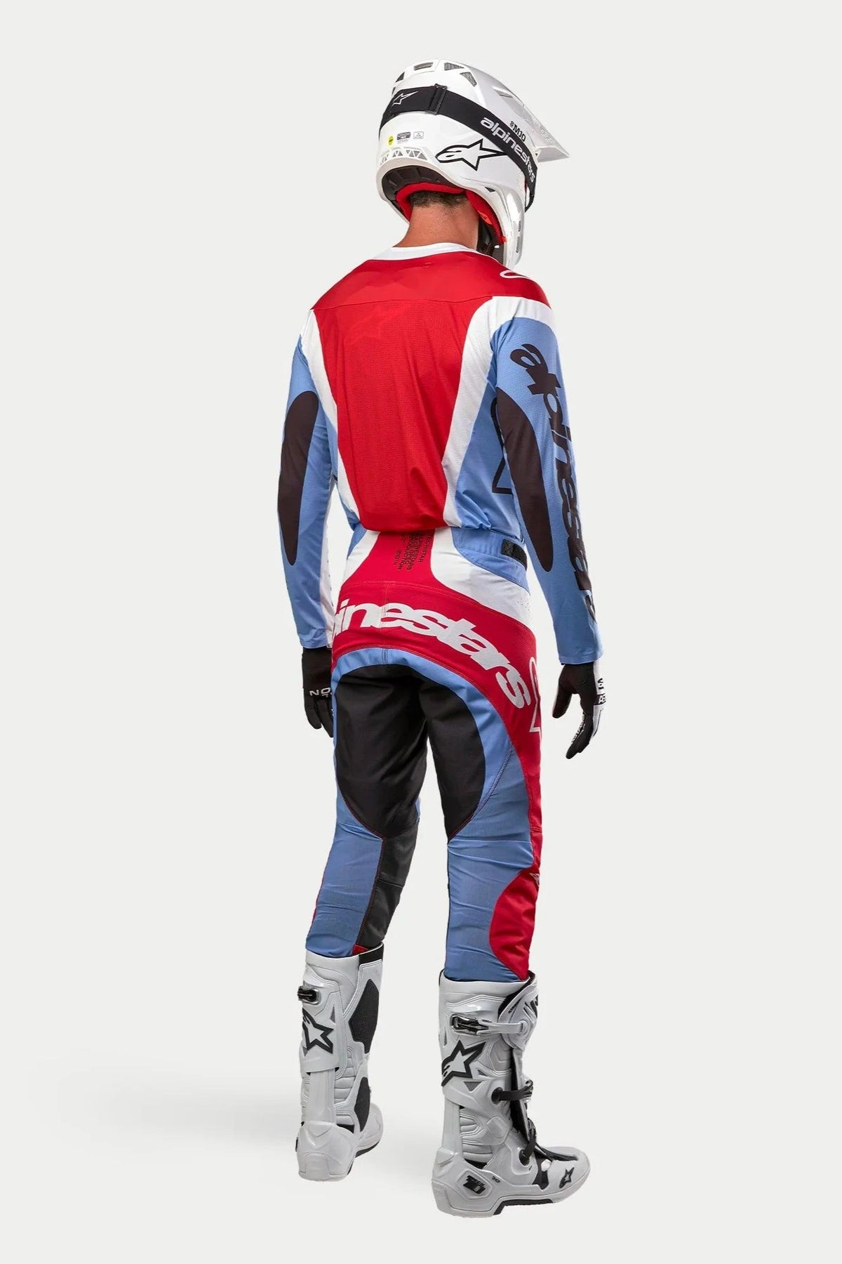 Motocross-rytter set bagfra, komplet racerudstyr, 2024 Techstar Ocuri Jersey i lyseblå, Mars-rød og hvid, matchende bukser med markant branding, hvide og sorte støvler, hvid hjelm, koordineret outfit til terrænkørsel