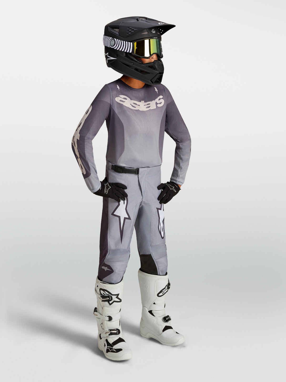 Alpinestars Ungdom Fluid Amber Gear Set, gråt motocrosssæt med grå trøje, bukser, hvide motorcykelstøvler og sort hjelm.