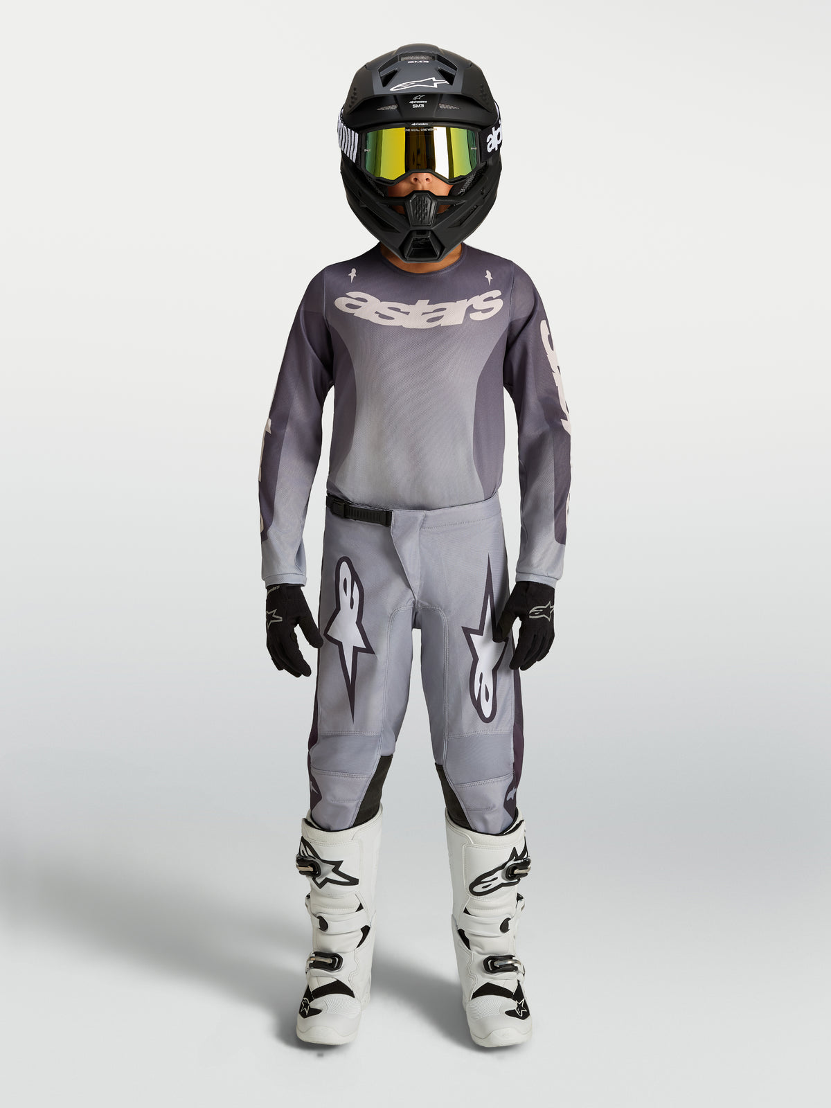 Alpinestars 2027 Ungdom Fluid Amber udstyrssæt, gråt og sandfarvet motocross-sæt med sort hjelm og hvide støvler.