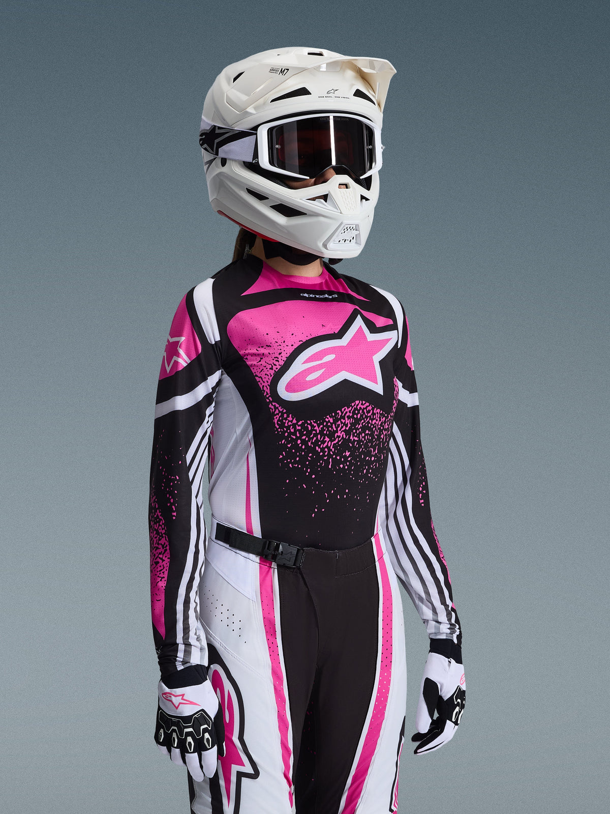 Female motocross rider wearing the Alpinestars 2026 Stella Techstar Nomur Gear Set in pink, black, and white; features a jersey with a large pink star logo, matching pants with magnetic closure buckle, a white helmet with goggles, and white and black gloves.