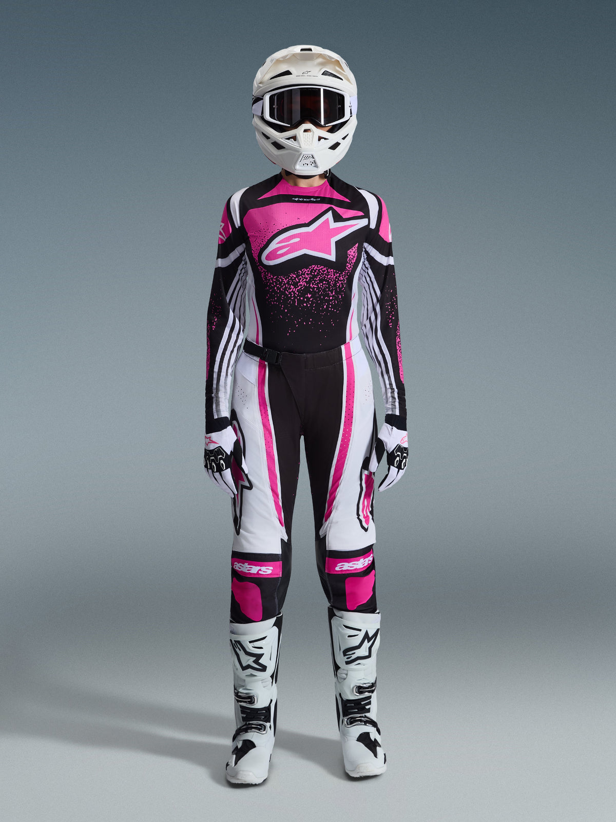 Female motocross rider wearing the Alpinestars 2026 Stella Techstar Nomur Gear Set, Pink, Black, and White racing kit, jersey featuring a large pink star logo and speckled graphic design, matching performance pants with white and pink panels, white helmet and goggles, white and black gloves, and white motocross boots