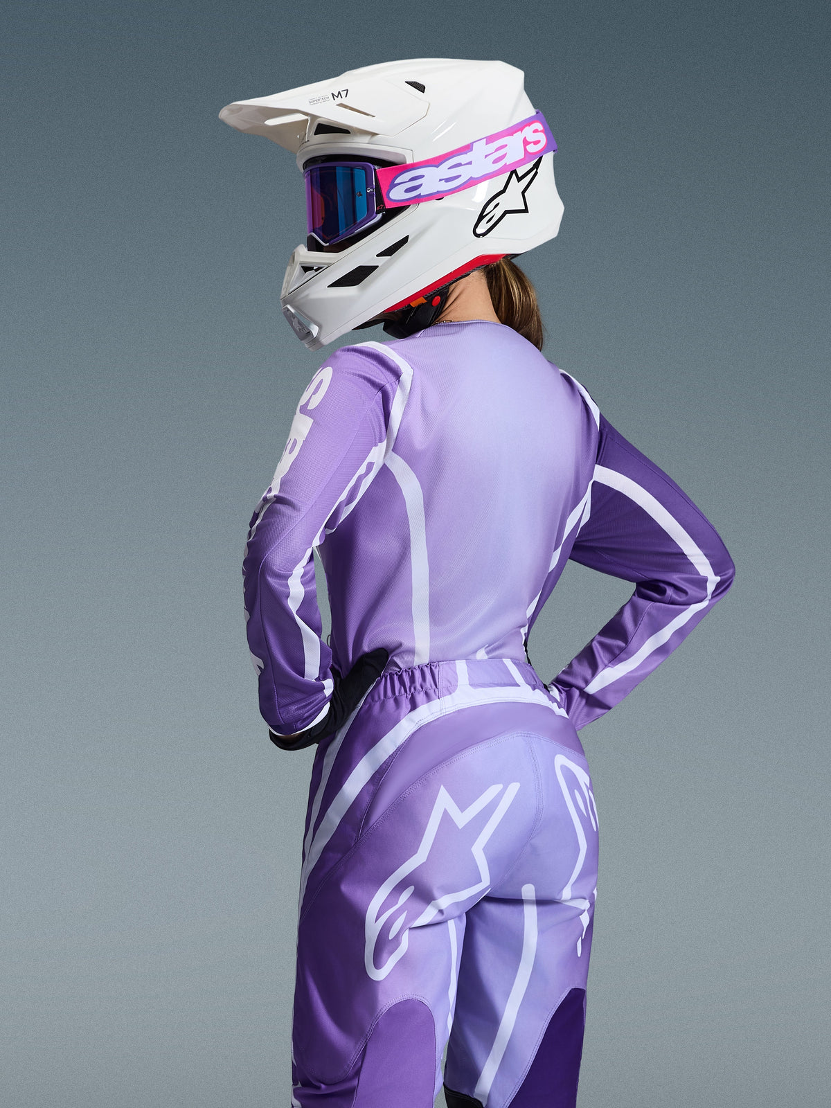 Alpinestars 2026 Stella Fluid Apex-udstyrssæt, motocross-kører iført lilla og lavendelfarvet trøje og bukser med hvid grafik, hvid hjelm med lyserøde og lilla goggles, specifik pasform til kvinder, koordineret udstyr til off-road-kørsel