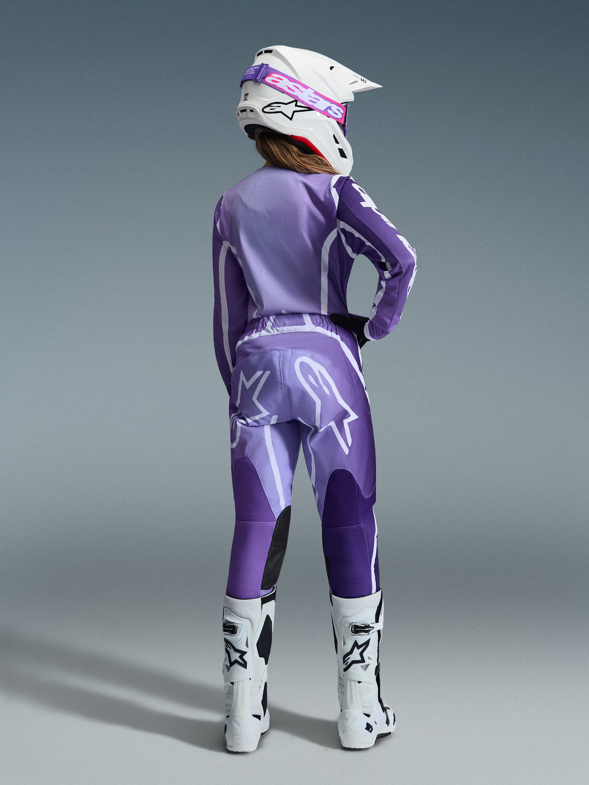 Alpinestars 2026 Stella Fluid Apex Gear Set, motocross-udstyr til kvinder, lilla og lavendel med hvide detaljer, kvindelig kører set bagfra iført matchende trøje og bukser, hvid hjelm med pink goggles og hvide støvler, koordineret atletisk raceroutfit