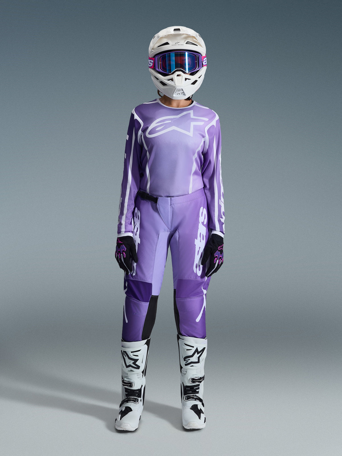 Motocross-kører iført Alpinestars 2026 Stella Fluid Apex-udstyrssæt, lilla trøje med hvidt logo, matchende lilla bukser med hvid branding, hvid hjelm med lyserøde goggles, hvide og sorte støvler, koordineret motocross-outfit til damer til terrænkørsel