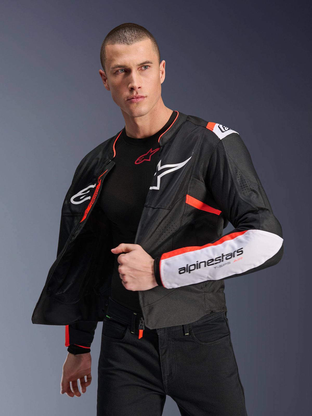 Male model wearing the Alpinestars T-SPS Air V2 Jacket, black textile motorcycle jacket with white and fluorescent red accents, featuring mesh panels for airflow, white Alpinestars logo on the chest and sleeve branding, worn unzipped over a black base layer