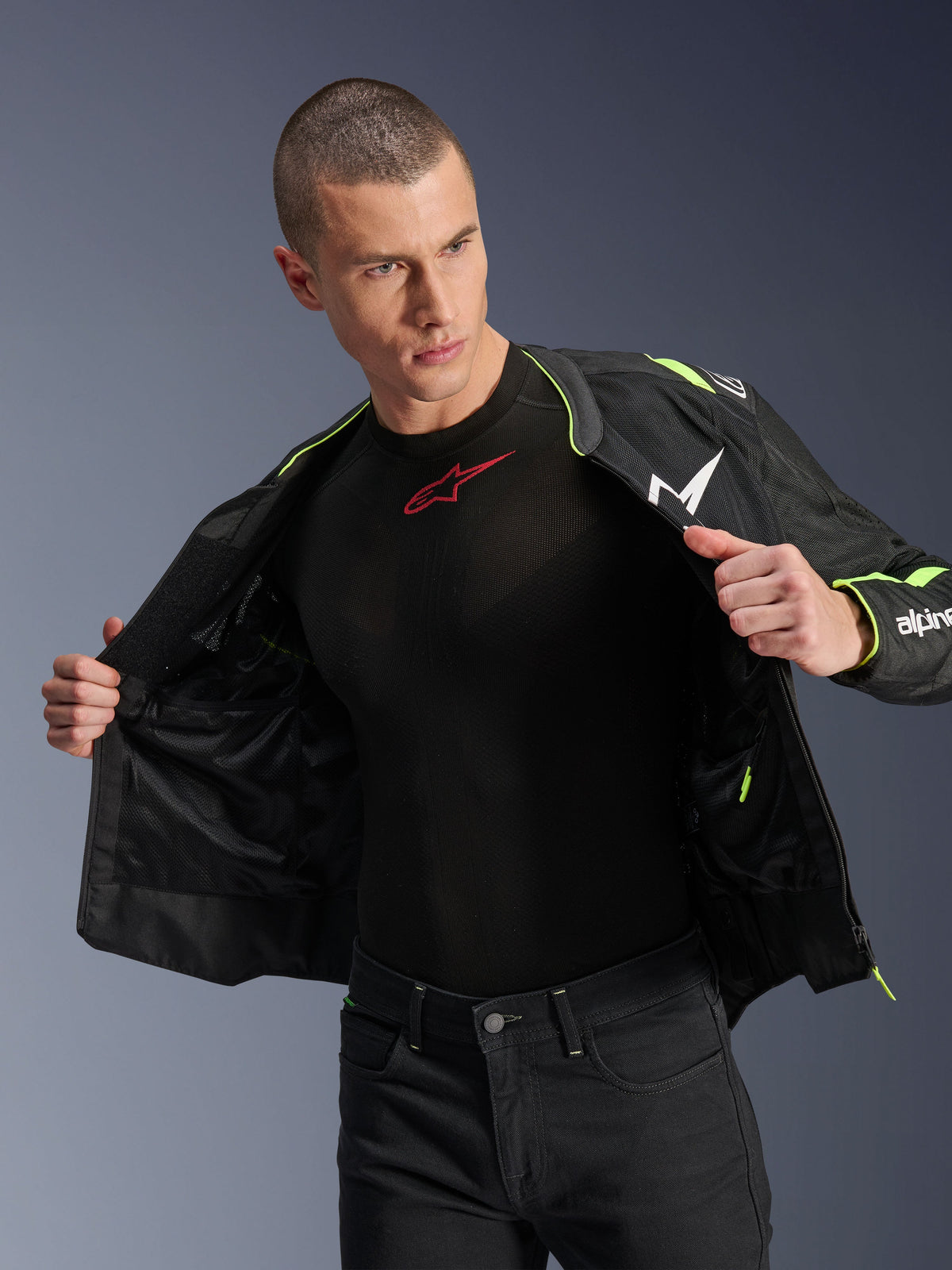 Mandlig model iført Alpinestars T-Sps Air v2-jakke i sort og gul fluo, vist åben for at fremhæve den indvendige mesh-konstruktion og lommer, kombineret med et sort Alpinestars baselayer og sorte bukser, urban motorcykeludstyr-stil