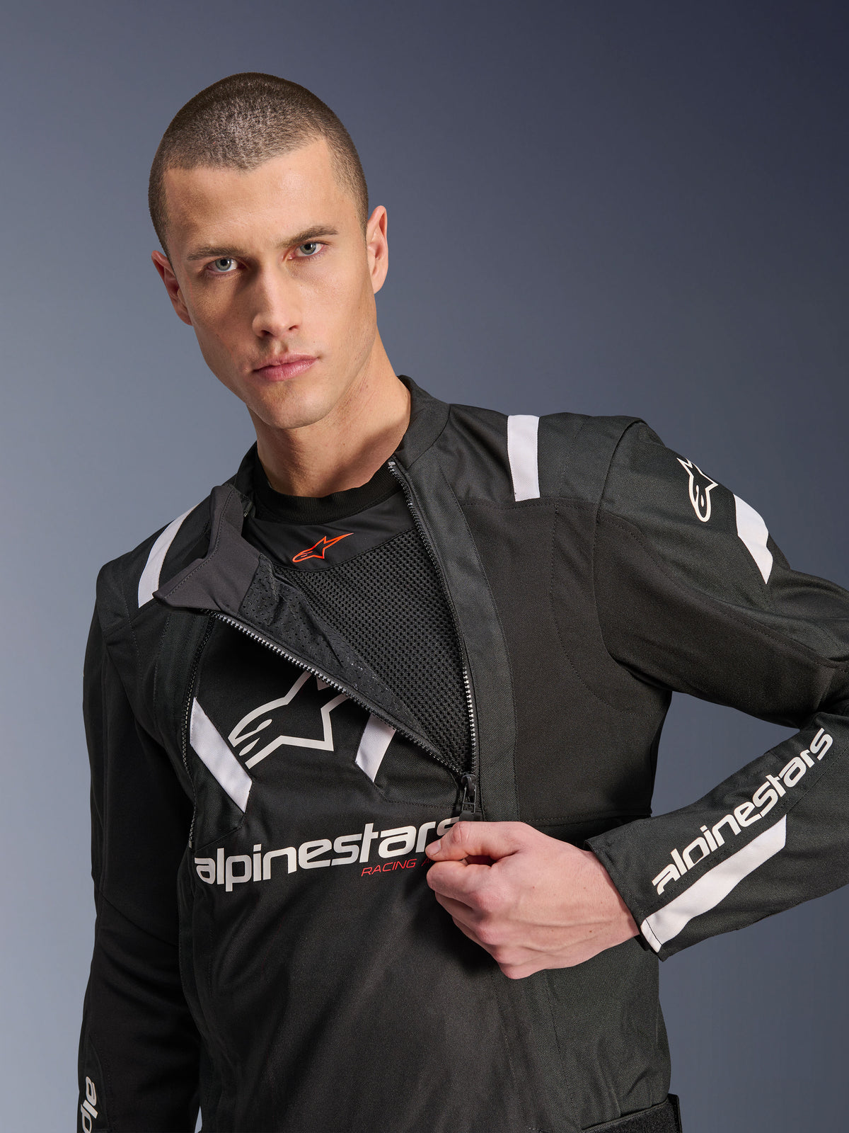 Alpinestars T-Stunt Air Jakke, sort og hvid motorcykeljakke, mandlig model viser delvist lynet modulært frontpanel der afslører mesh-lag til luftstrøm, hvide Alpinestars-logoer på bryst og ærmer, sportsligt design til landevejskørsel