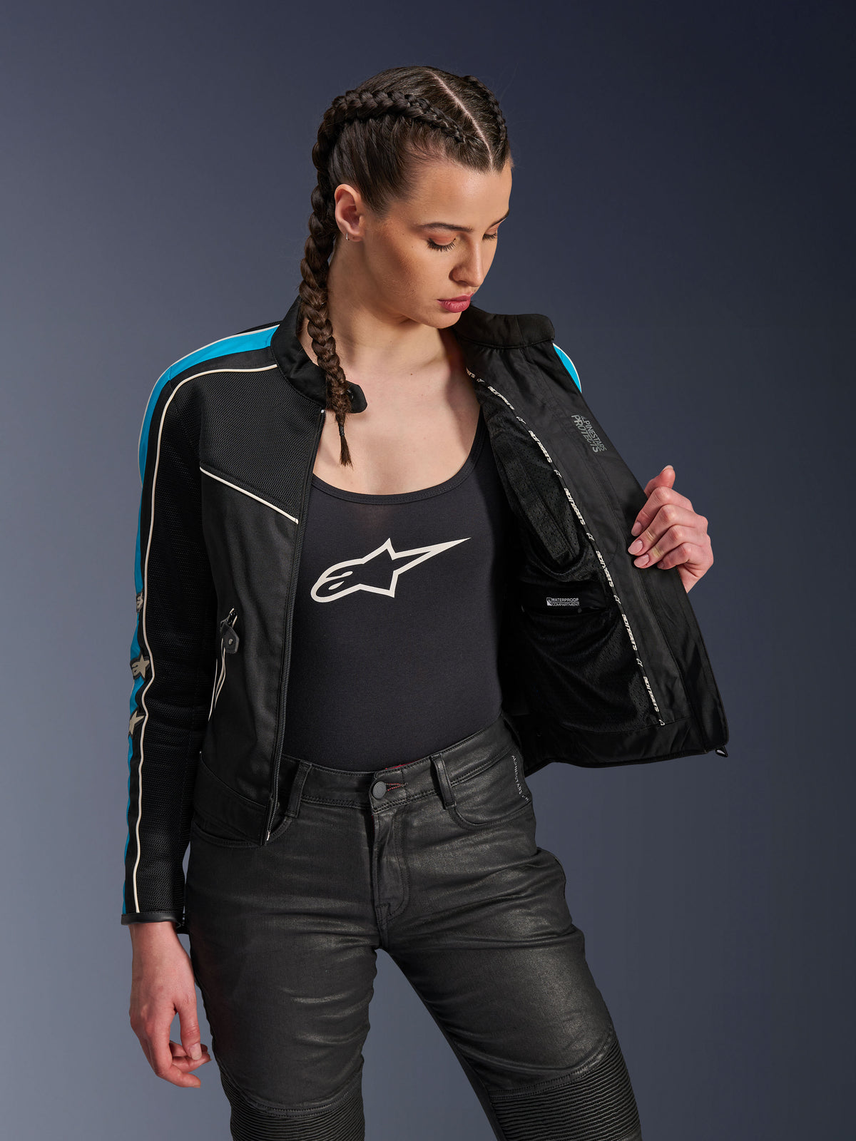 Kvinde iført Alpinestars Stella T-Dyno Air-jakken i Black Light Blue Ecru, der holder jakken åben for at vise det indvendige mesh-for og den vandtætte lomme, kombineret med en sort tanktop med logo og sorte kørebukser, vintage-inspireret urbant motorcykeludstyr.