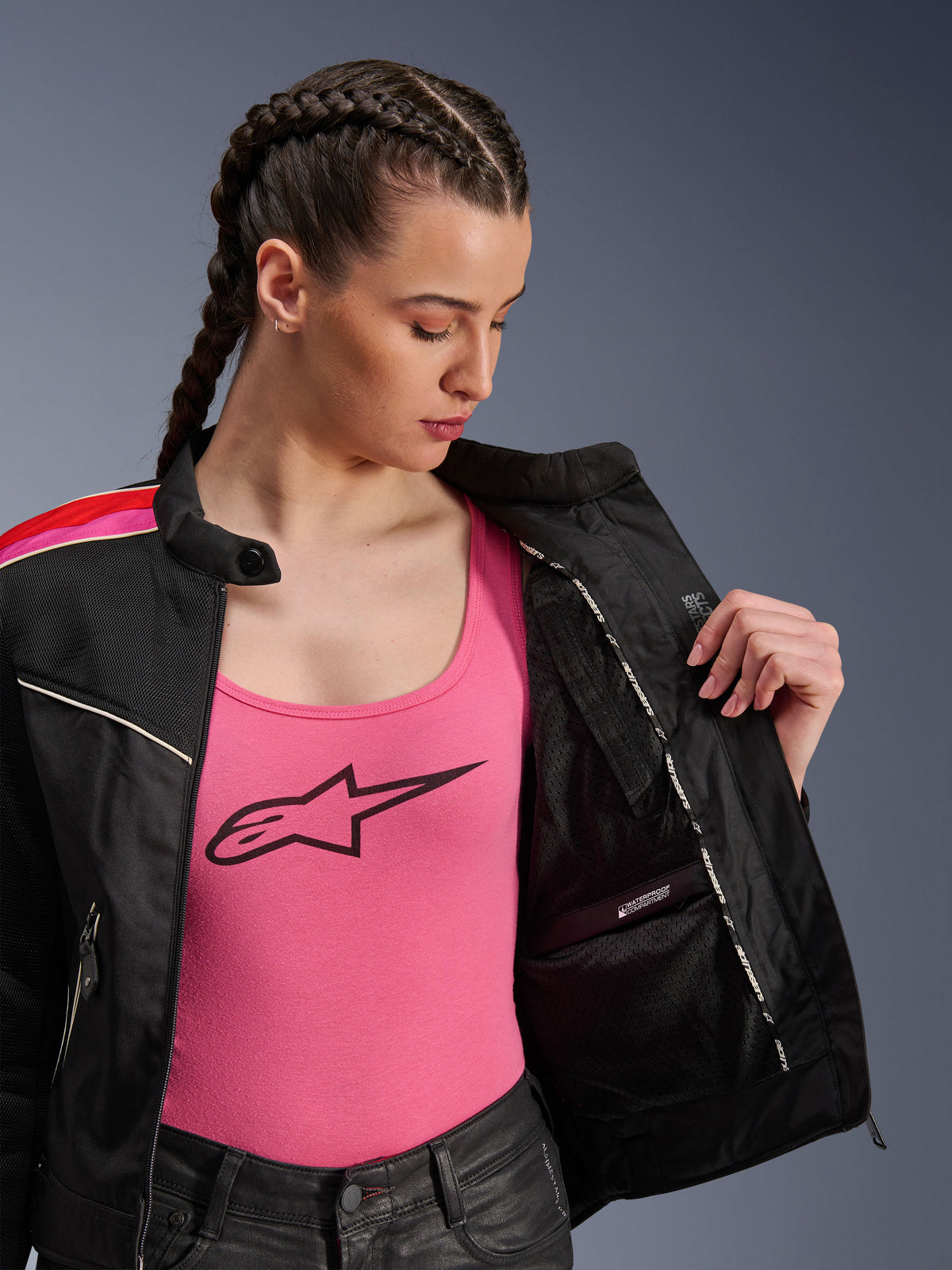 Kvindelig motorcyklist iført Alpinestars Stella T-Dyno Air-jakken i Black Bright Red Fuchsia, vist delvist åben for at afsløre det indvendige vandtætte rum, båret over en lyserød tanktop med logo, hvilket fremhæver de åndbare mesh-paneler og den skræddersyede bikerpasform.