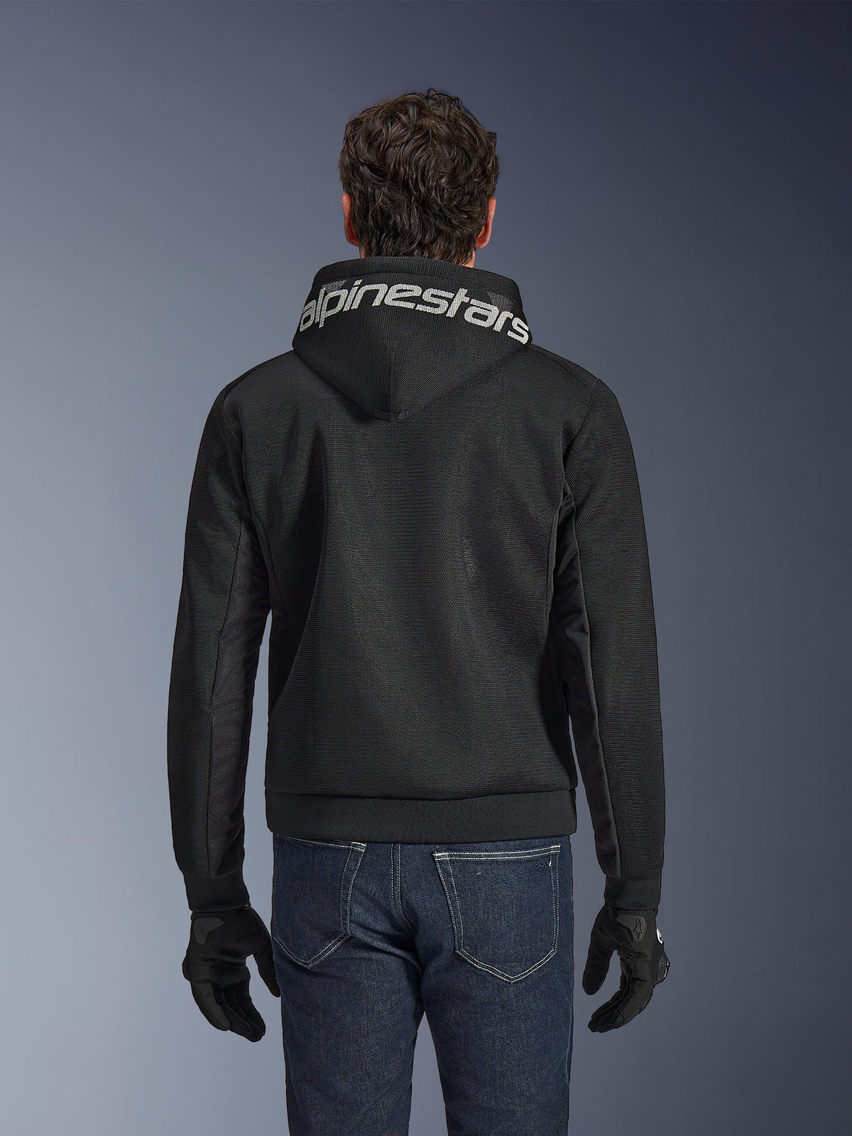 Alpinestars Chrome Superairflow Hoodie, sort motorcykelhættetrøje, set bagfra med markant hvid Alpinestars-branding på hætten, 3D teknisk mesh-konstruktion, bæres med mørkeblå jeans og sorte handsker, ventileret beklædning til bykørsel