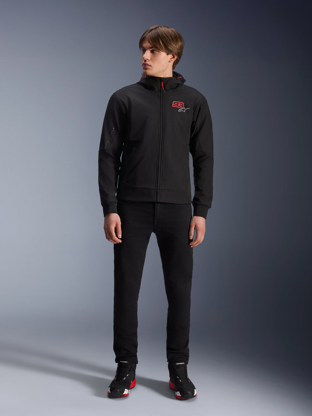 Alpinestars MM93 Radium Tech Hoodie, sort urban motorcykelhættetrøje, rødt 93-logo og hvidt stjernelogo på brystet, båret af en model med sorte bukser og matchende køresko, strømlinet vindtæt teknisk shell til bykørsel