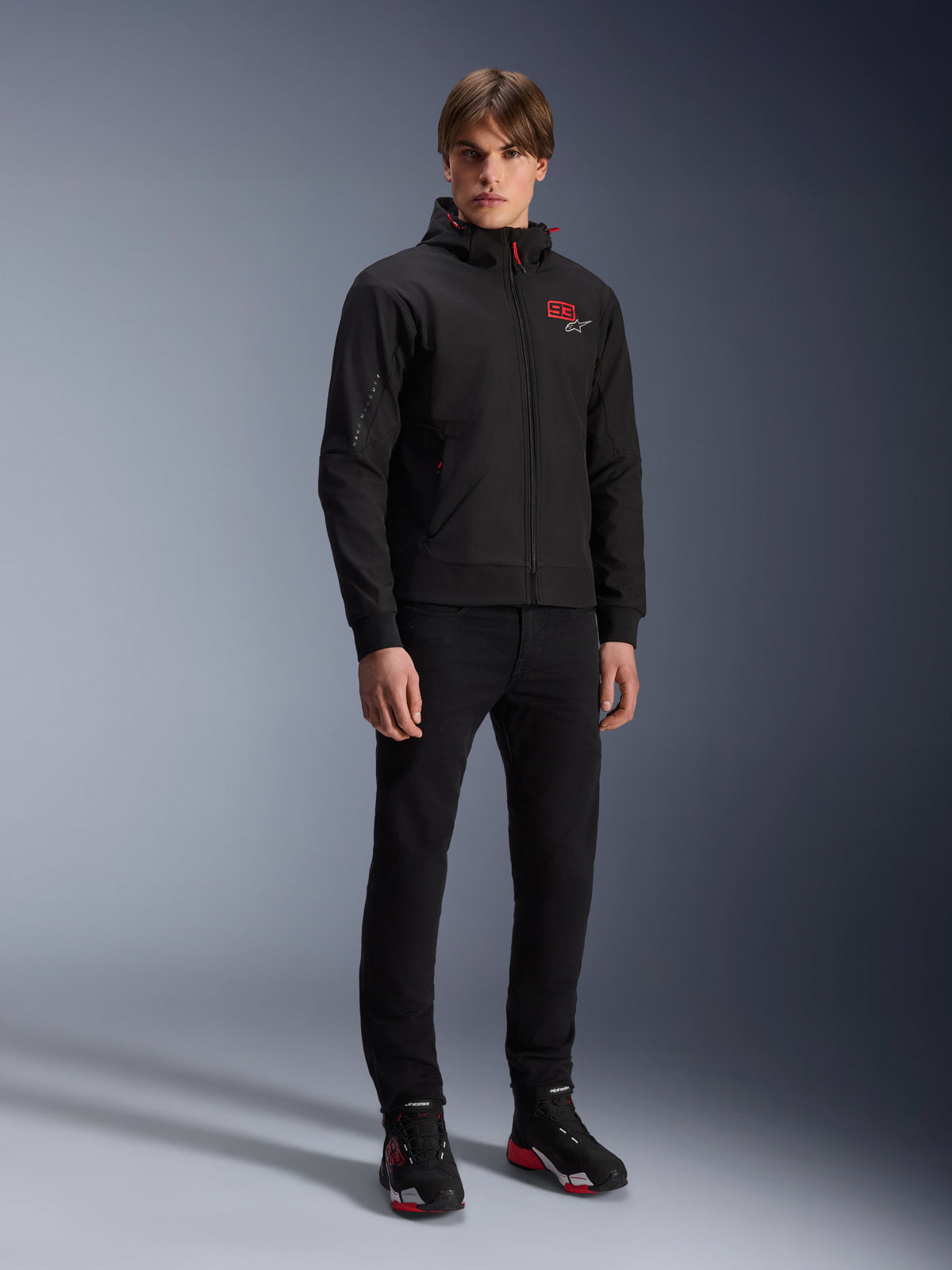 Alpinestars MM93 Radium Tech Hoodie, urban motorcykeljakke, sort med klare røde detaljer, båret af model med sorte bukser og køresko, med rødt '93'-logo på brystet og Marc Marquez-branding på ærmet, afslappet teknisk streetwear til bykørsel