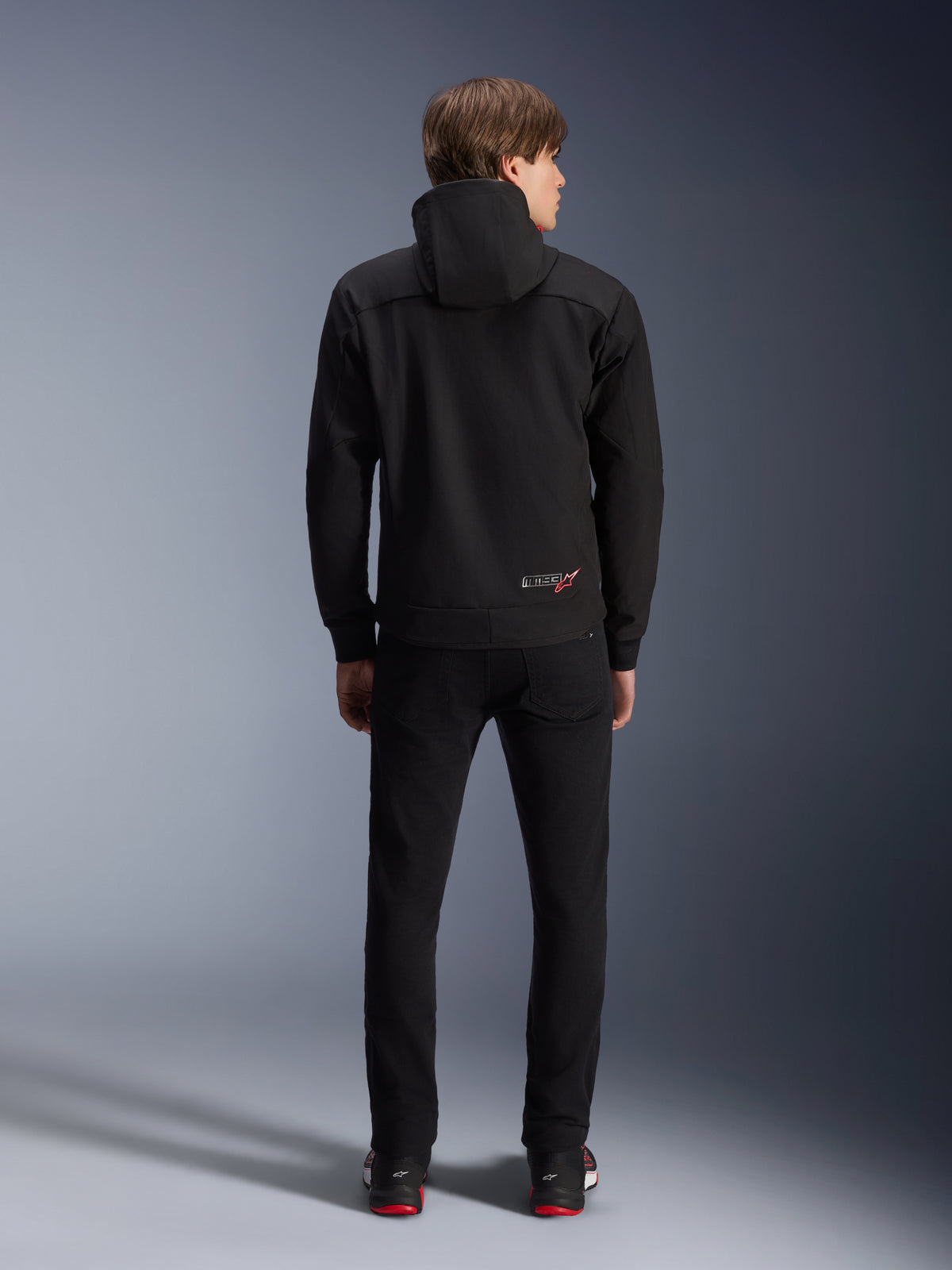 Alpinestars MM93 Radium Tech Hoodie, sort, set bagfra på model, med et lille MM93-logo med rød stjerne nederst på ryggen, kombineret med sorte bukser, urban motorcykelbeklædning