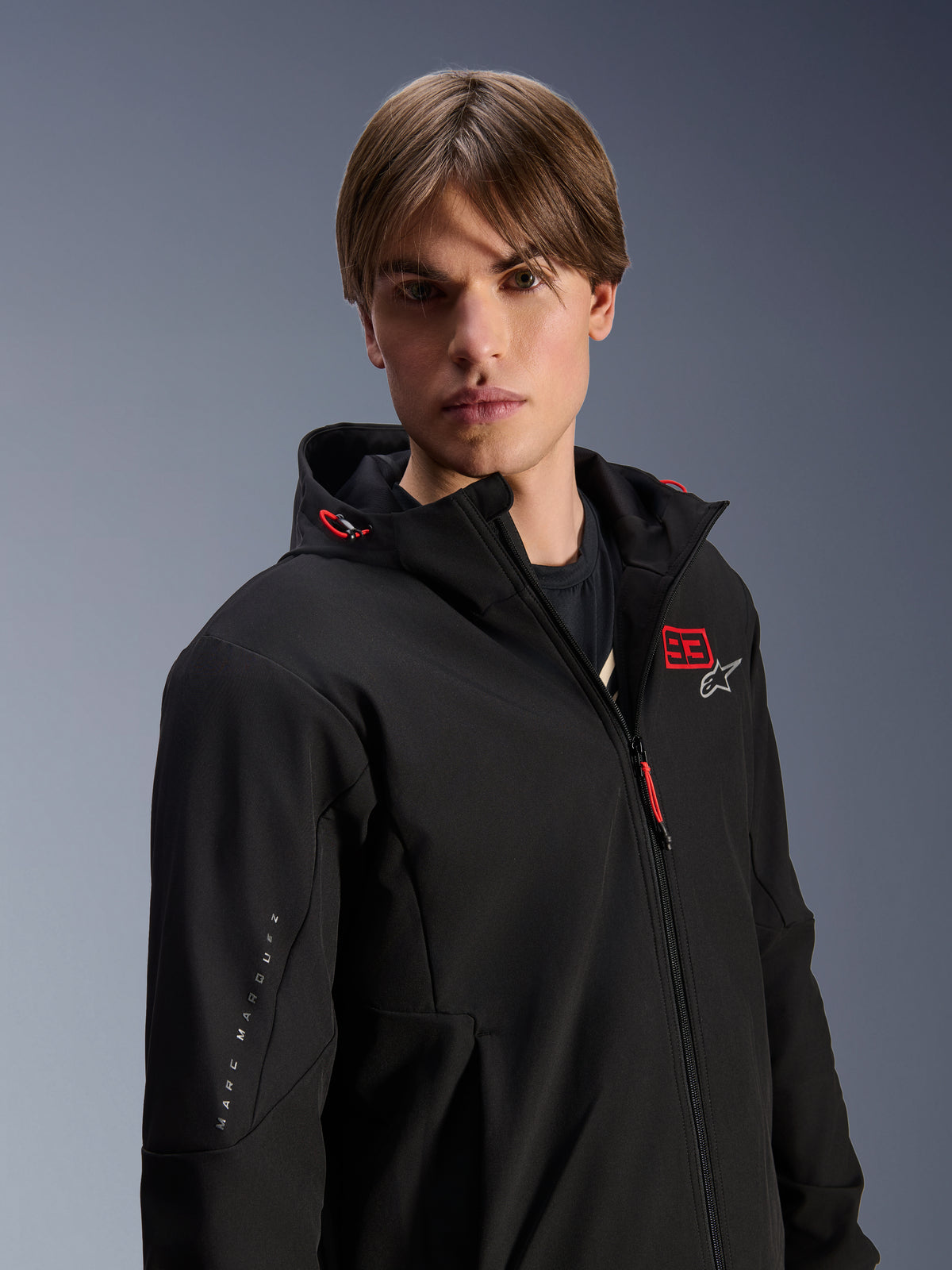 Mandlig model iført Alpinestars MM93 Radium Tech Hoodie, sort urban motorcykeljakke med rødt '93' og hvidt stjernelogo på brystet, 'Marc Marquez' branding på ærmet, teknisk vindtæt skal til bykørsel.