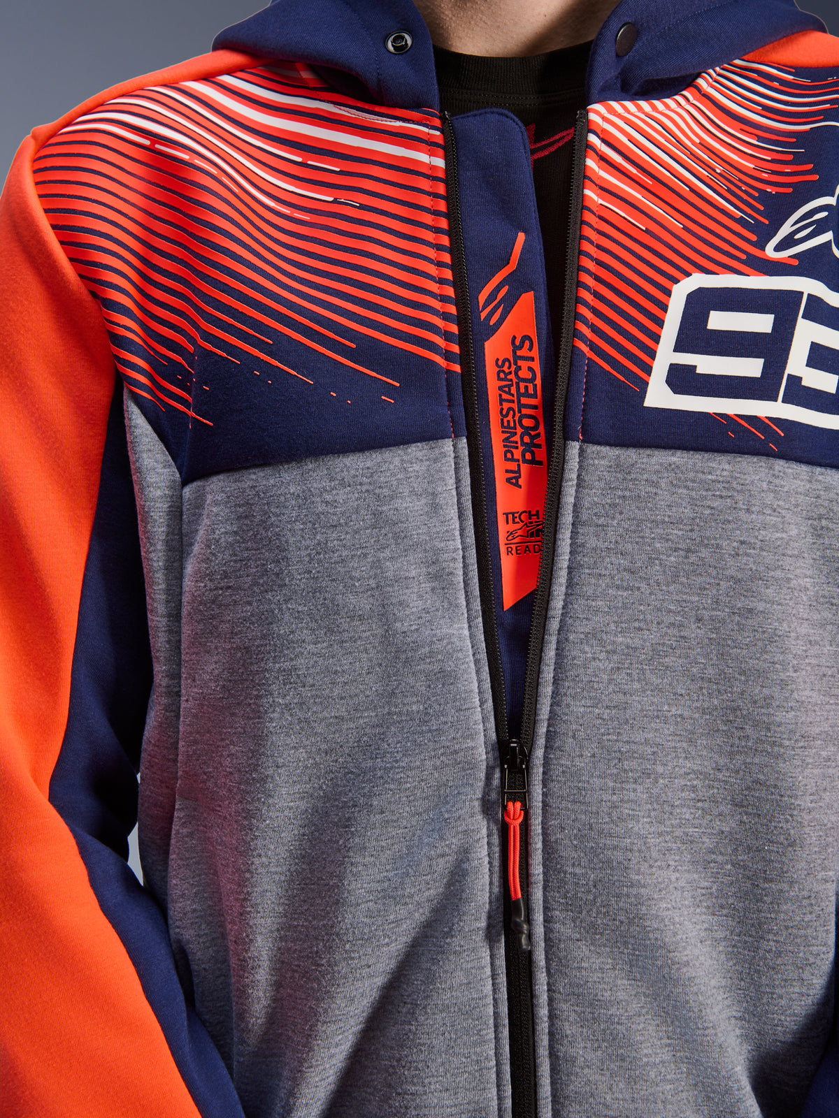 Alpinestars MM93 Chrome V2 Sport Hoodie, close-up of chest and collar, navy blue and gray melange tech fleece with red fluo graphics, featuring the white 93 logo and Alpinestars Protects branding on the inner zipper flap, Tech-Air Ready motorcycle riding apparel.