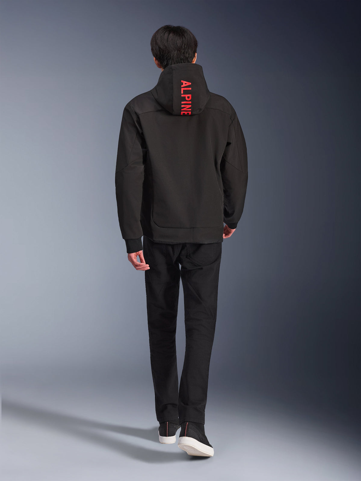 Alpinestars Radium Tech Hoodie, urban motorcykeljakke, sort, set bagfra med klar rød 'ALPINE'-tekst på hætten, kombineret med sorte bukser og sneakers, strømlinet teknisk skal til pendling.