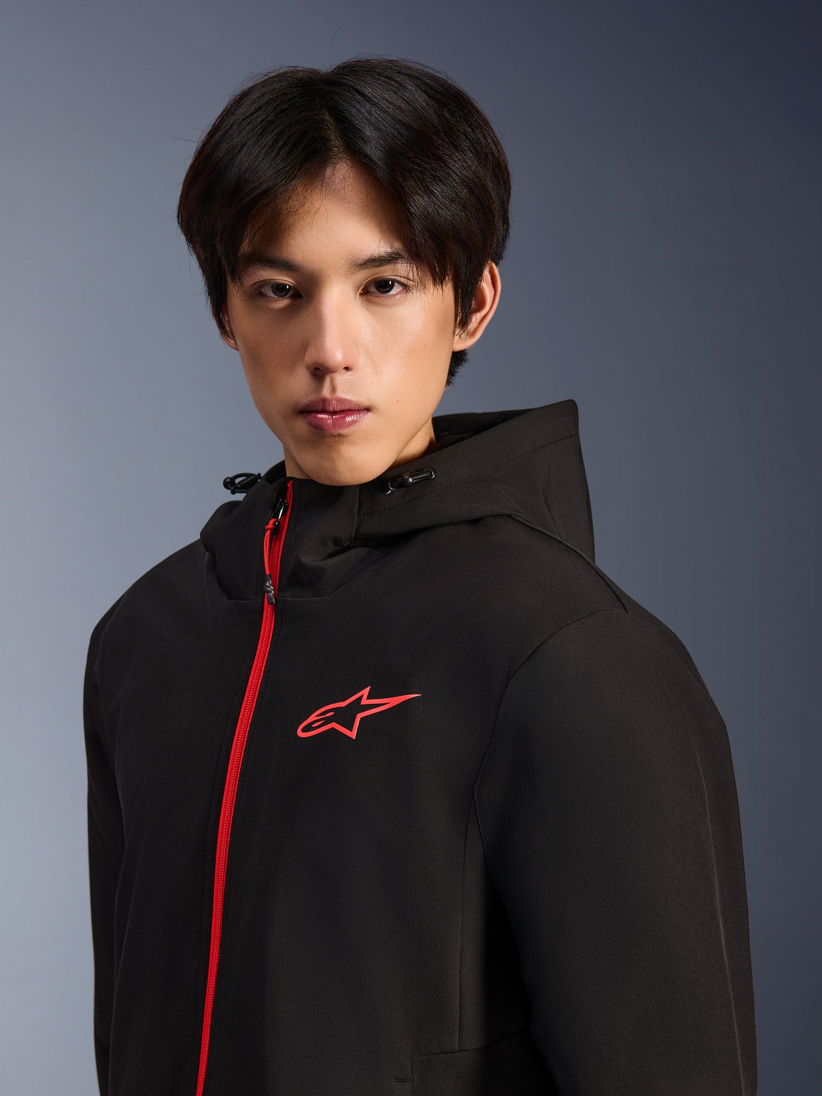 Alpinestars Radium Tech Hoodie, urban motorcykeljakke, sort og lysende rød, mandlig model iført en teknisk shell-hættetrøje med en lysende rød lynlås foran og brystlogo, moderne urbant køredesign