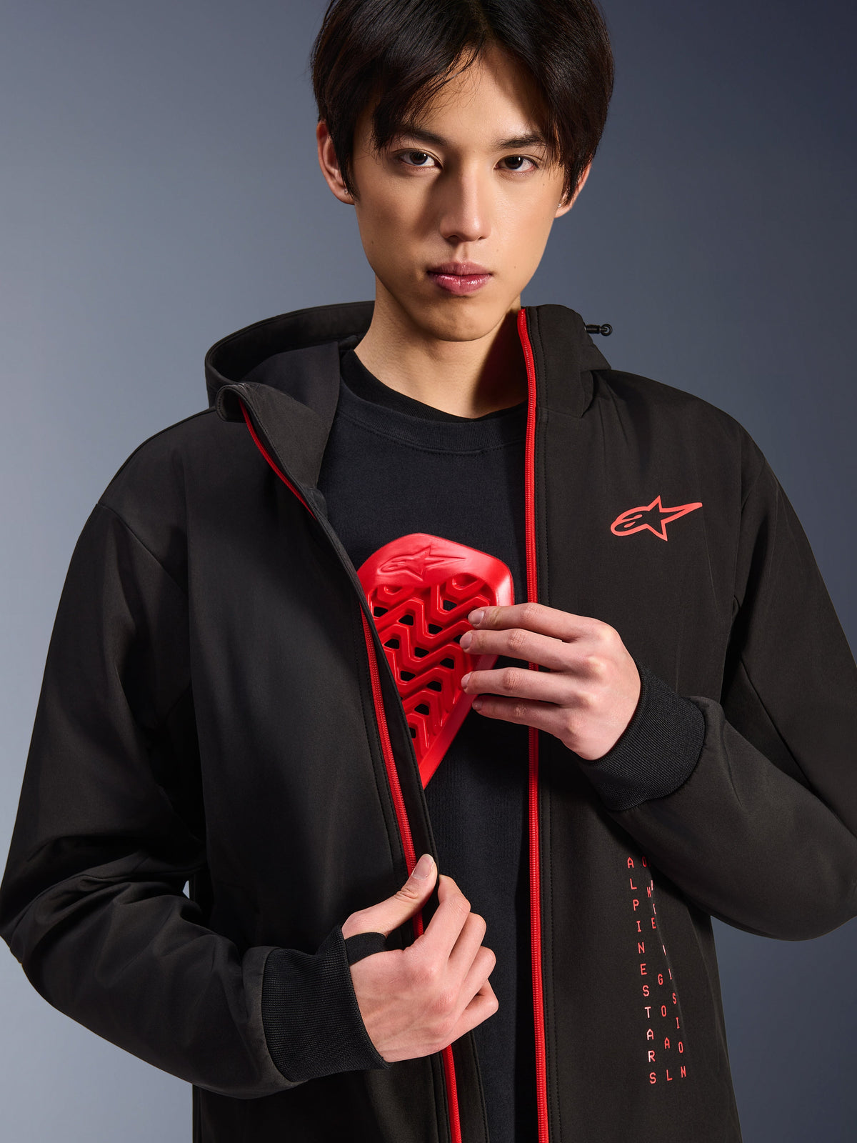 Alpinestars Radium Tech Hoodie, urban motorcykeljakke, sort og stærk rød, båret af en model, der viser et rødt brystpanser-indlæg, har stærkt røde lynlåse og lodret Alpinestars-branding, strømlinet vindtæt teknisk skal til pendling i byen