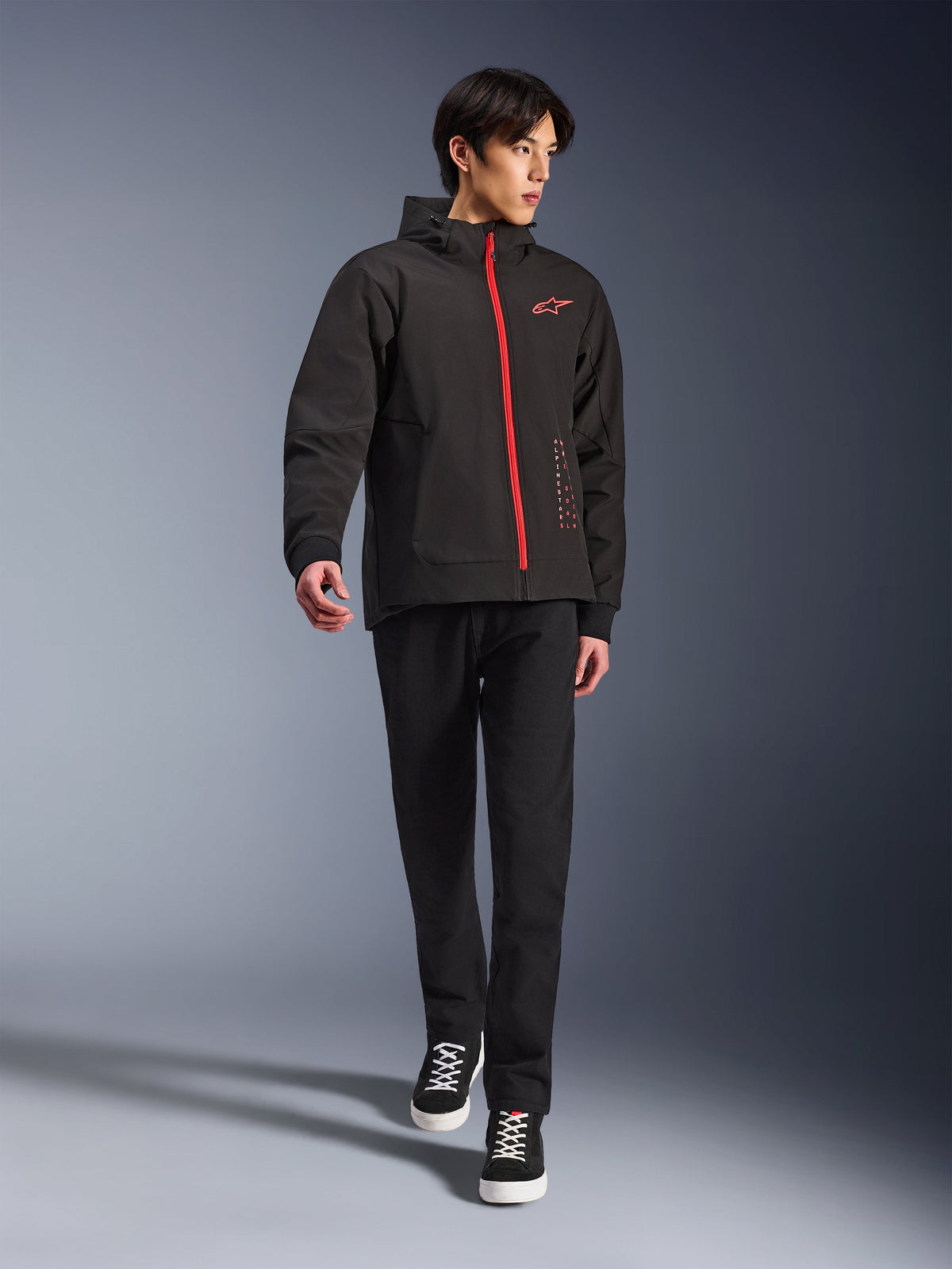 Alpinestars Radium Tech Hoodie i Black Bright Red, urban motorcykeljakke båret af en model, med rød lynlås foran og logo på brystet, stylet med sorte bukser og sneakers for et moderne pendlingslook.