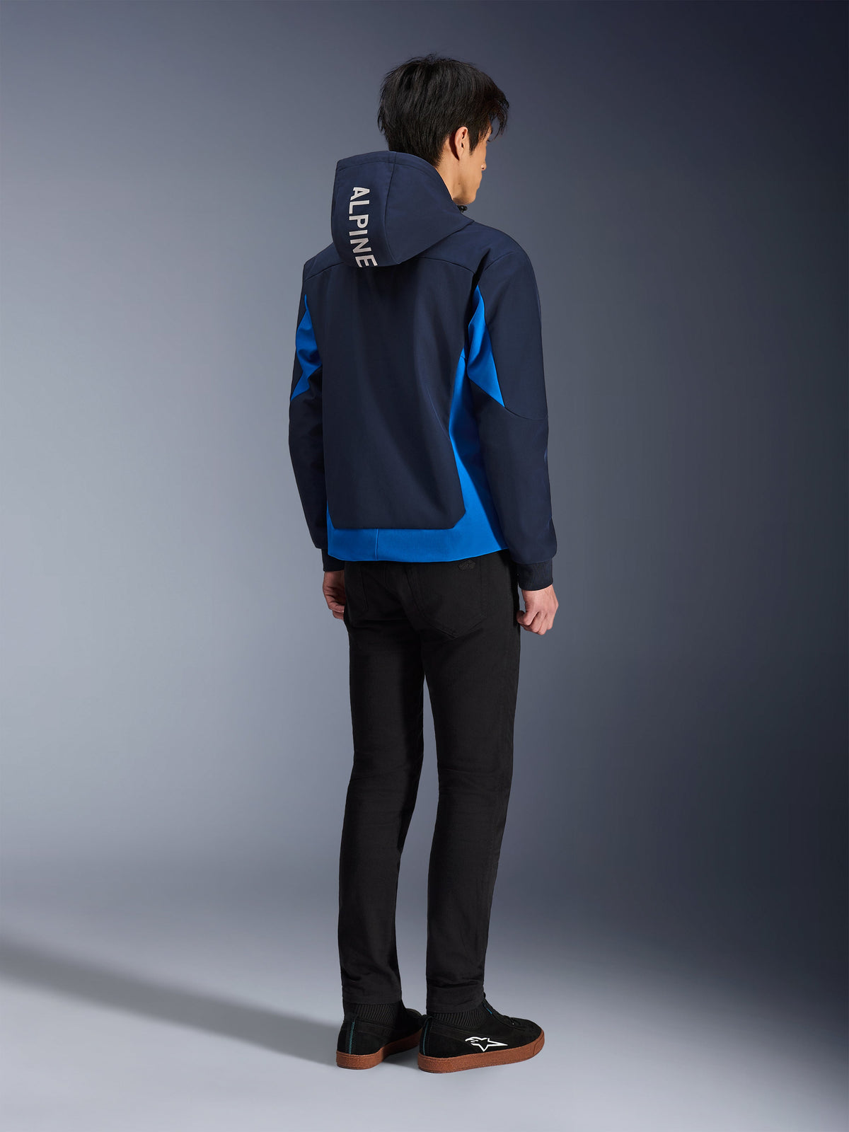 Alpinestars Radium Tech Hoodie, Night Navy og Royal Blue, set bagfra af model iført den tekniske shell-hættetrøje med hvid 'ALPINE' tekst på hætten, parret med sorte bukser og sko, urbant motorcykeludstyr