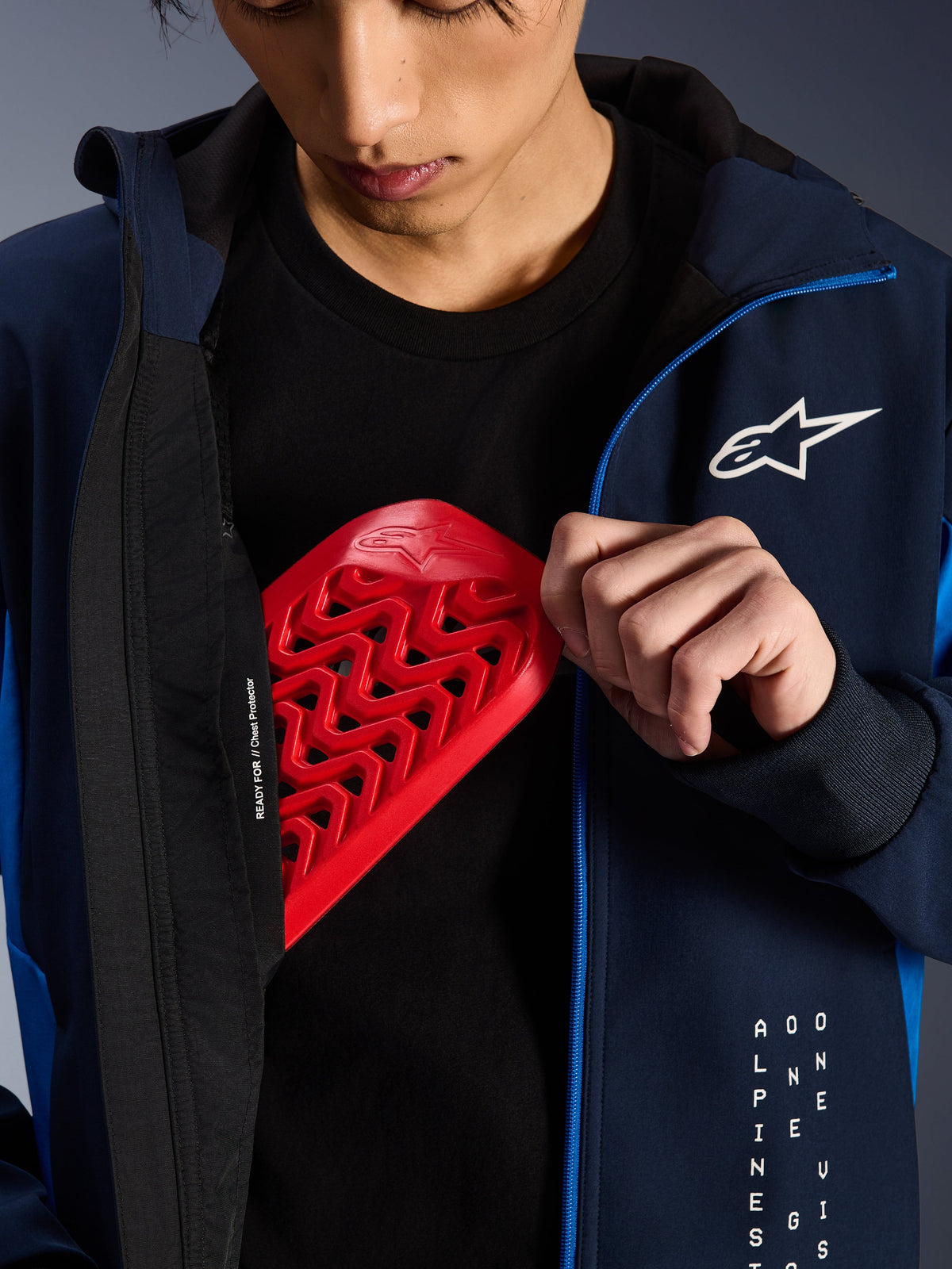 Alpinestars Radium Tech Hoodie i Night Navy og Royal Blue, nærbillede der viser en rød Nucleon brystbeskytter blive placeret i den indvendige lomme, med 'READY FOR // Chest Protector' tekst på foret og et hvidt stjernelogo på brystet.