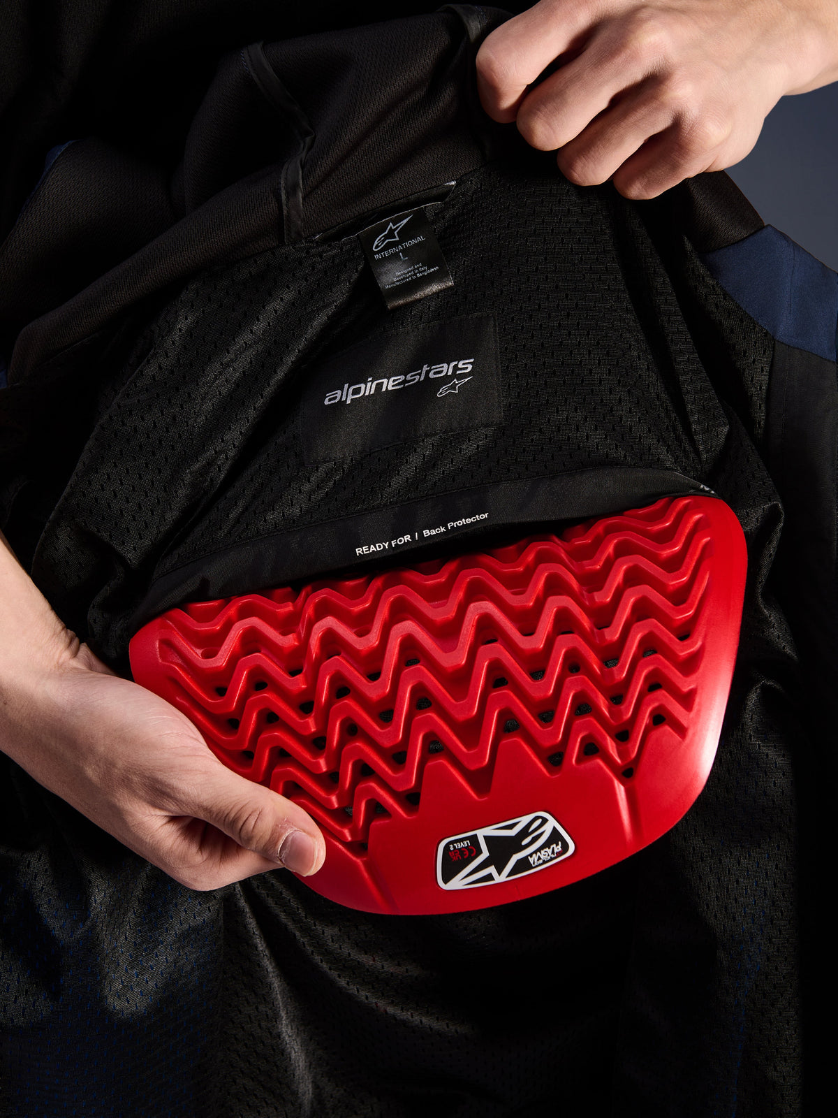 Alpinestars Radium Tech Hoodie indvendig detalje, rød Nucleon Plasma rygbeskytter bliver sat ind i rygbeskytterlommen, sort mesh-for med 'READY FOR | Back Protector' tekst, bølget honeycomb-design, tekniske beskyttelsesfunktioner til urban motorcykeljakke