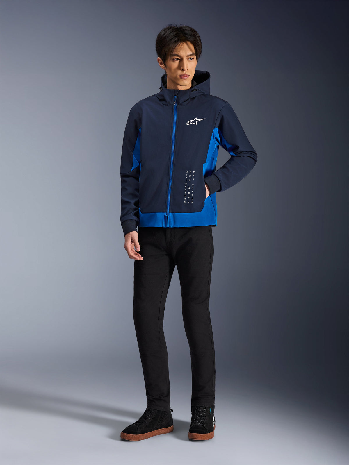 Alpinestars Radium Tech Hoodie, urban motorcykeljakke, Night Navy og Royal Blue, båret med sorte bukser og sorte sneakers, med et hvidt Alpinestars-logo på brystet og lodret tekstdetalje, afslappet atletisk pasform til bykørsel