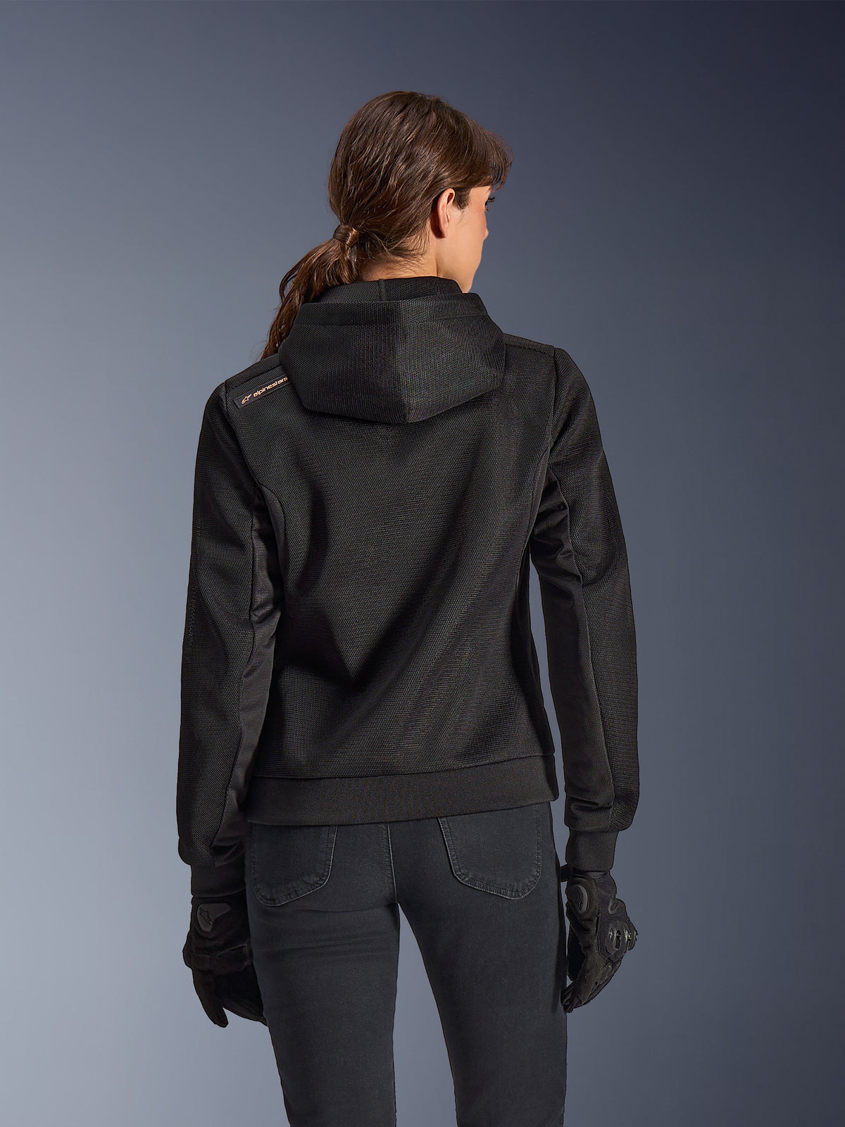 Alpinestars Stella Chrome Superairflow Hoodie, set bagfra af kvinde iført sort teknisk 3D-mesh motorcykelhættetrøje, har konstrueret hætte og kobberfarvet logo på skulderen, kombineret med sorte motorcykelhandsker og bukser.