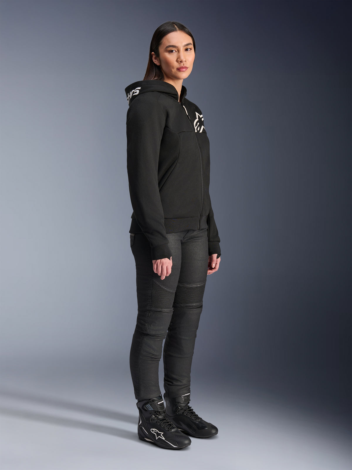 Kvinde iført Alpinestars Stella Chrome V2 Sport hættetrøje i sort, med hvidt Alpinestars-logo, kombineret med sorte motorcykelbukser og støvler, urbant køreudstyr til kvinder med en afslappet atletisk pasform