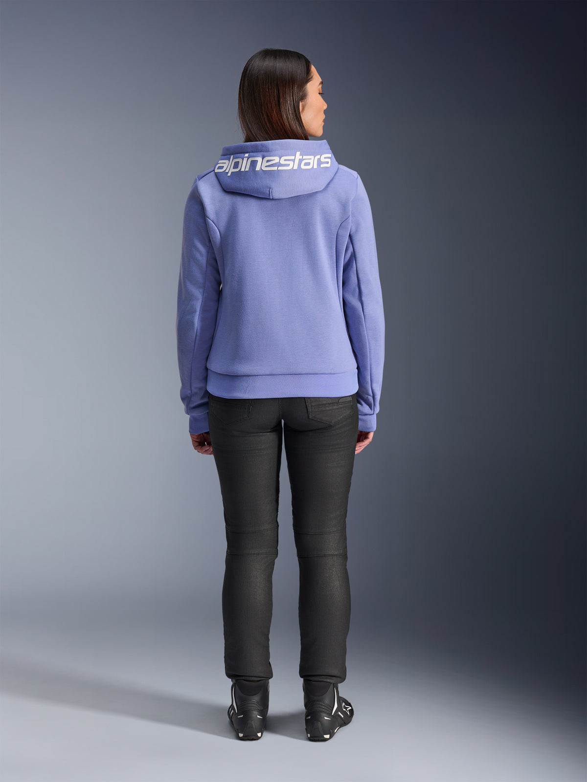 Alpinestars Stella Chrome V2 Sport Hoodie, Blue Violet, set bagfra af en kvinde iført hættetrøjen med hvidt Alpinestars-logo på tværs af hætten, kombineret med sorte bukser og kørestøvler, afslappet urban motorcykelbeklædning