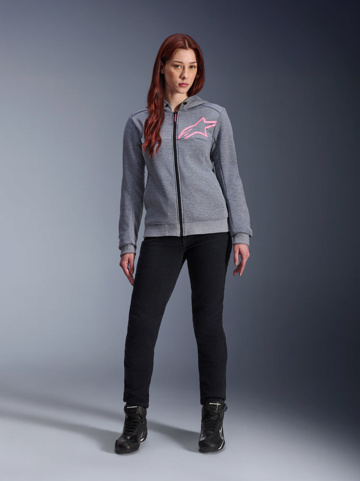 Alpinestars Stella Chrome V2 Sport Hoodie, Gray Heather Pink Fluo, båret af en kvinde med sorte bukser og støvler, med et lyserødt Alpinestars-stjernelogo på brystet, urbant motorcykeludstyr med gennemgående lynlås, letvægts tech-fleece med en afslappet atletisk pasform.