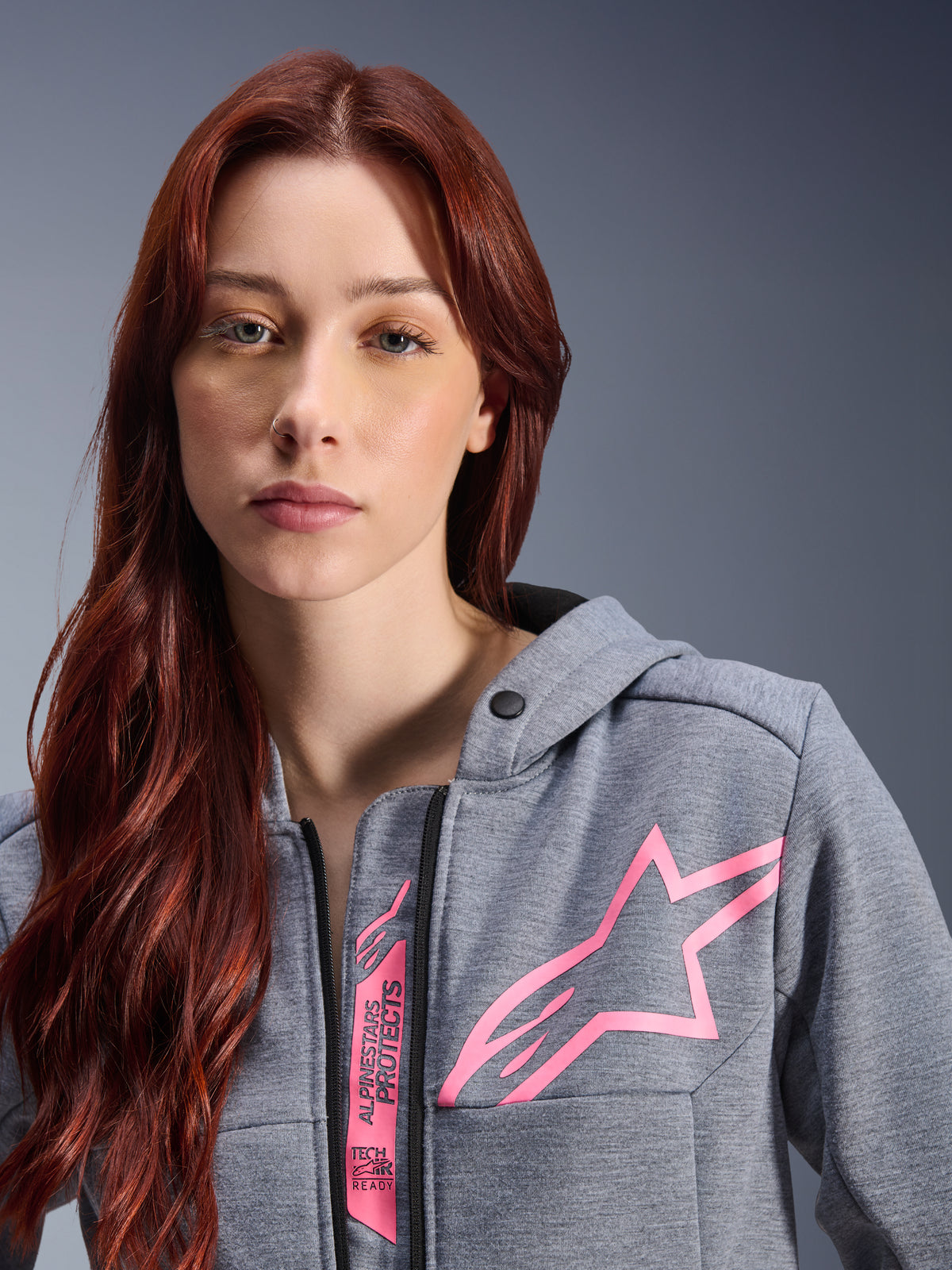 Kvinde iført Alpinestars Stella Chrome V2 Sport hættetrøje, Gray Heather Pink Fluo, nærbillede af det lyserøde Alpinestars stjernelogo og Tech-Air Ready-branding, urban motorcykelbeklædning